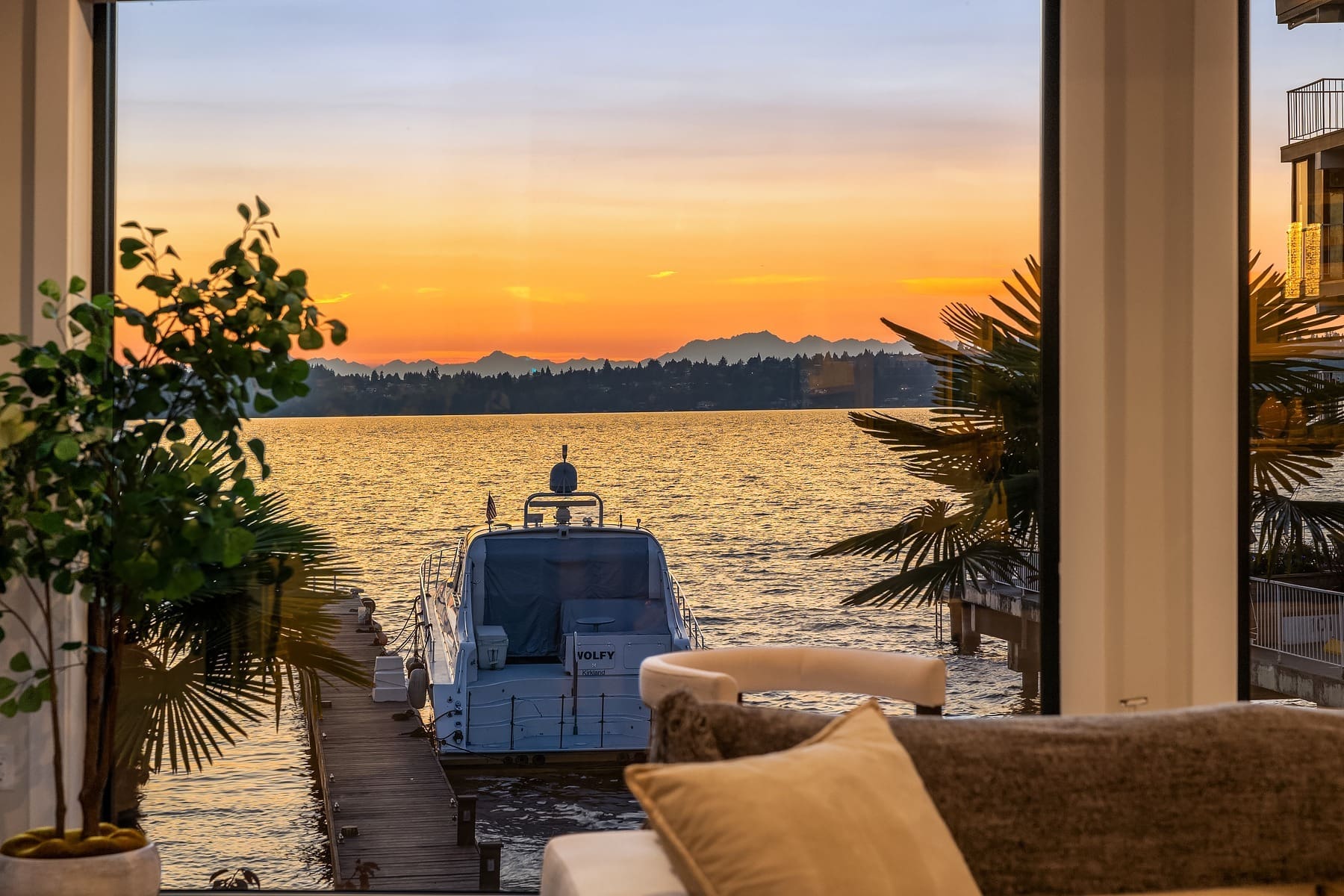 A luxurious interior with large windows offers a stunning view of a sunset over a calm lake. A docked yacht is seen outside, surrounded by greenery and neighboring buildings. The sky is painted in vibrant hues of orange and yellow.