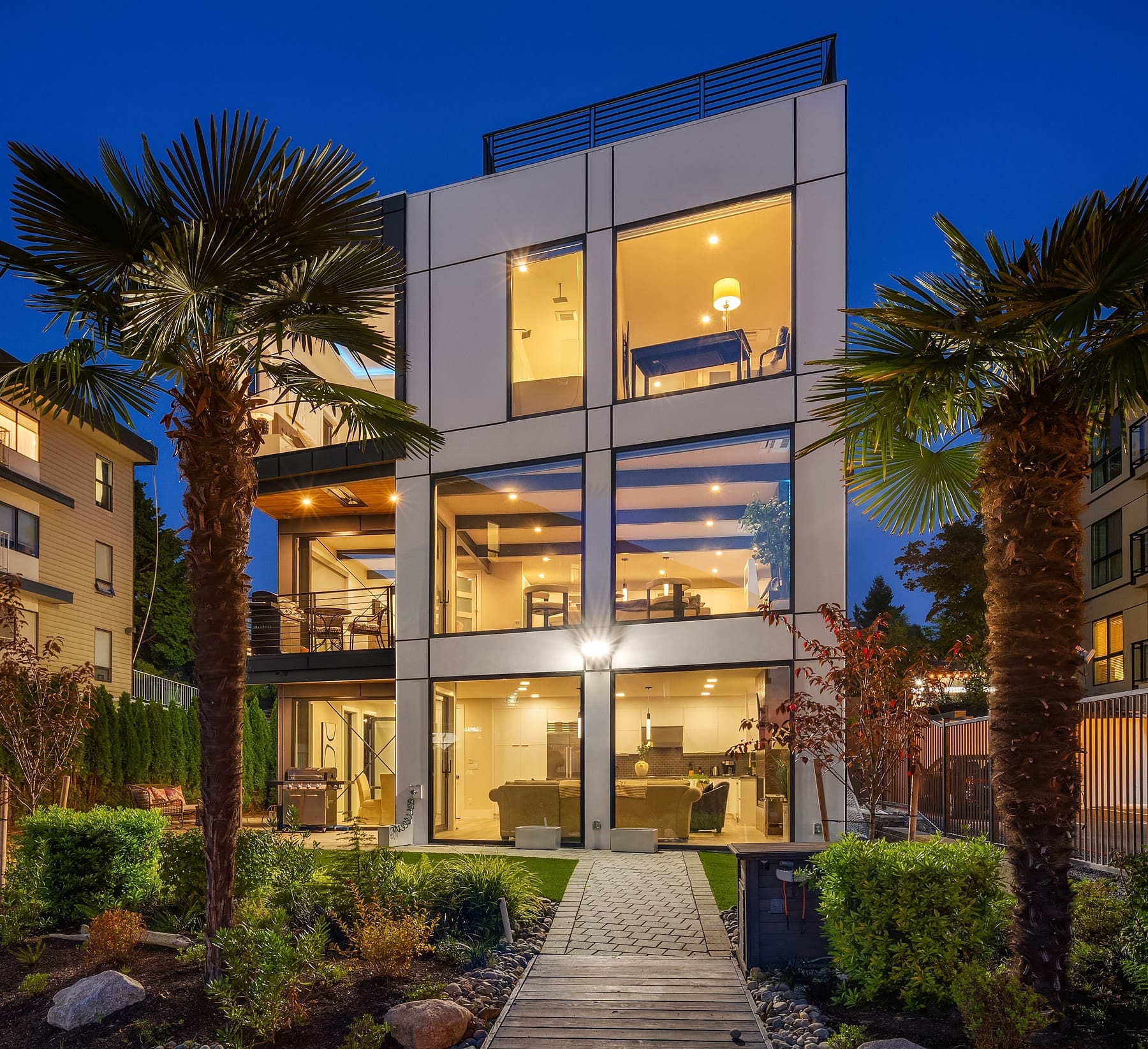 A modern three-story house with large floor-to-ceiling windows lit from within at night. The house has a minimalist design and is surrounded by a well-maintained garden, featuring palm trees and a stone pathway leading up to the entrance.