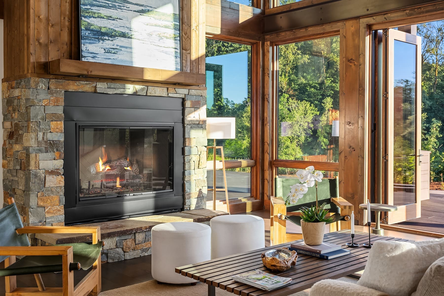 A cozy living room with a stone fireplace and a modern black insert. Large glass windows and doors let in ample natural light and offer a view of lush greenery outside. The room features comfortable seating, a wooden coffee table, and elegant décor.