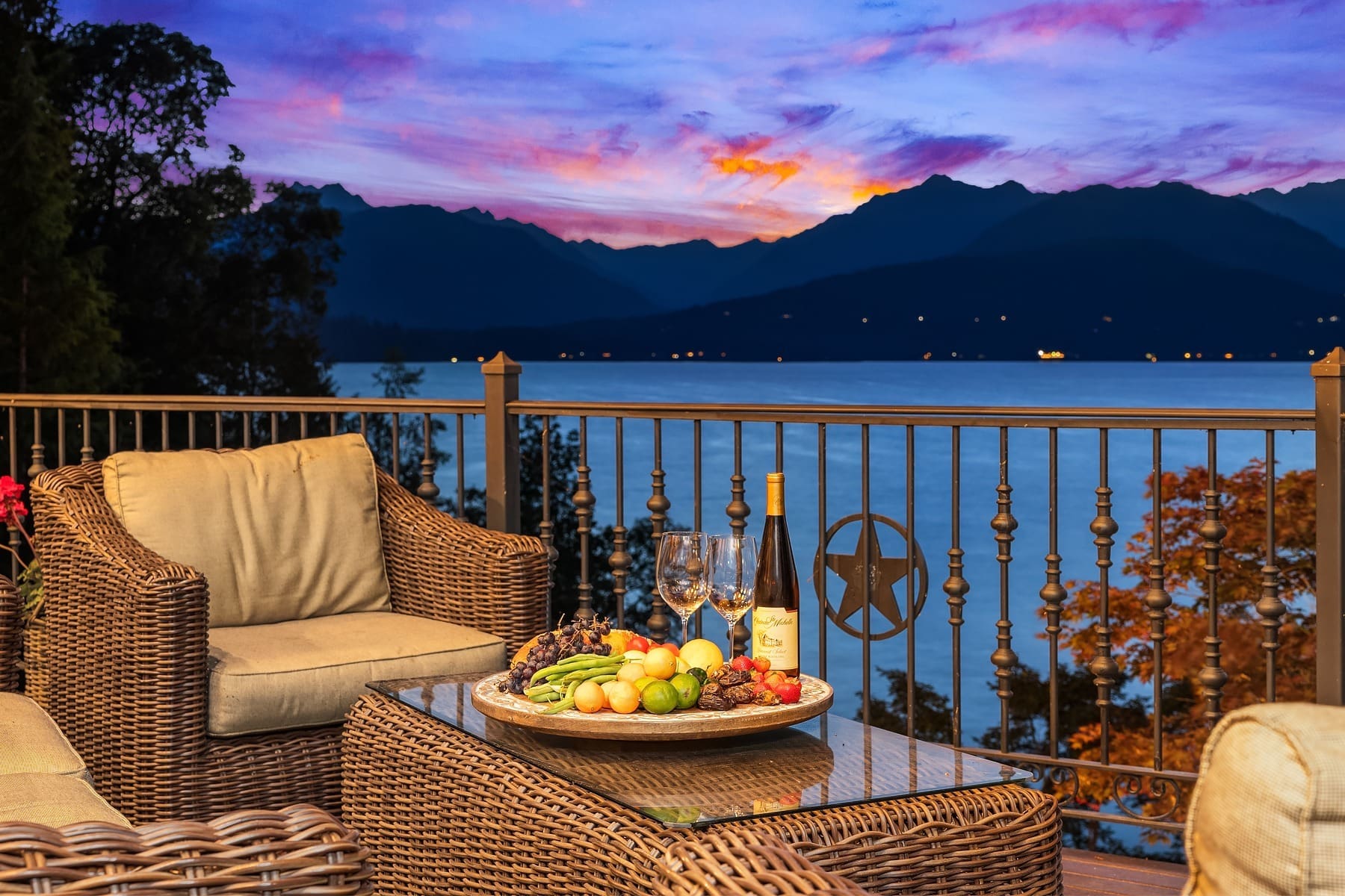 A cozy outdoor seating area with rattan furniture overlooking a serene lake and mountain range at sunset. A glass coffee table holds a wine bottle, two glasses, and a platter of assorted fruits. The sky is painted with vibrant hues of orange, pink, and purple. A charming, dark brown cottage with illuminated windows stands amidst a landscaped lawn at dusk. A small tree with a spotlight and an outdoor seating area enhance the cozy atmosphere. The clear sky overhead features a crescent moon, and tall trees frame the background. Twilight shot.