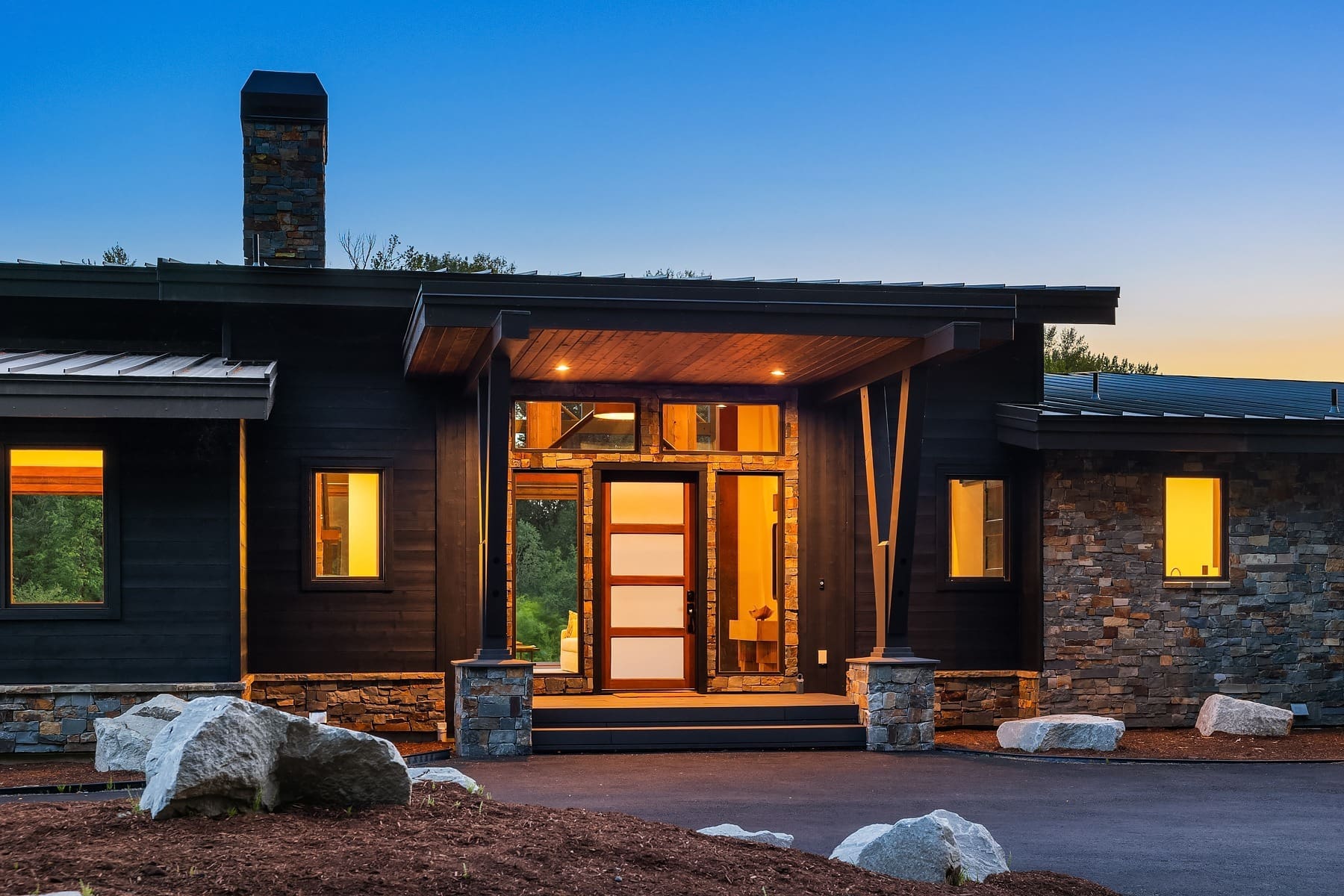 A modern single-story house with a flat roof, large windows, and a prominent front entrance. The house exterior features a combination of dark wood and stone, illuminated by warm lights. Large rocks and a landscaped front yard add a natural touch to the scene.