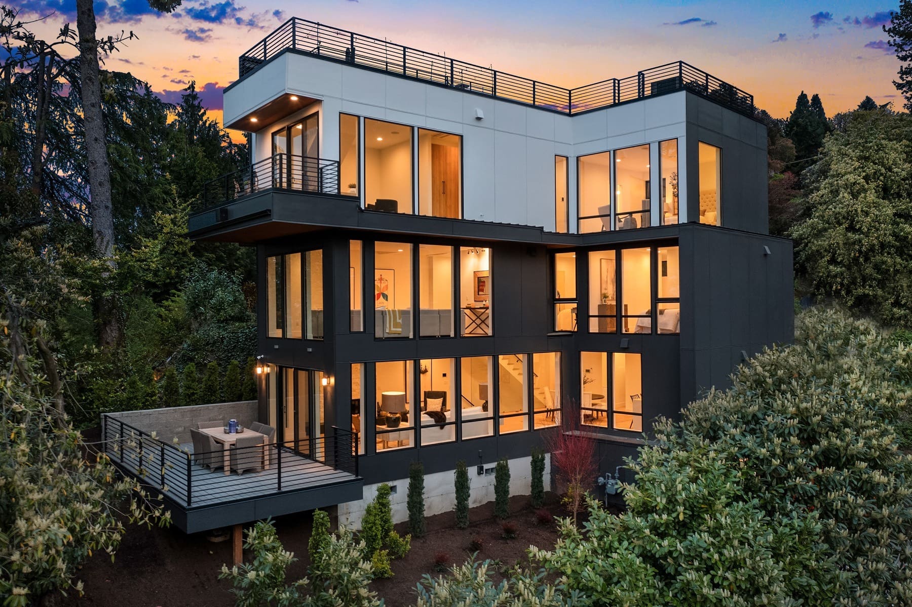 A modern, three-story house with large glass windows and multiple balconies. The house is surrounded by lush greenery, and the sky in the background is tinted with the colors of a sunset, creating a warm and inviting ambiance.