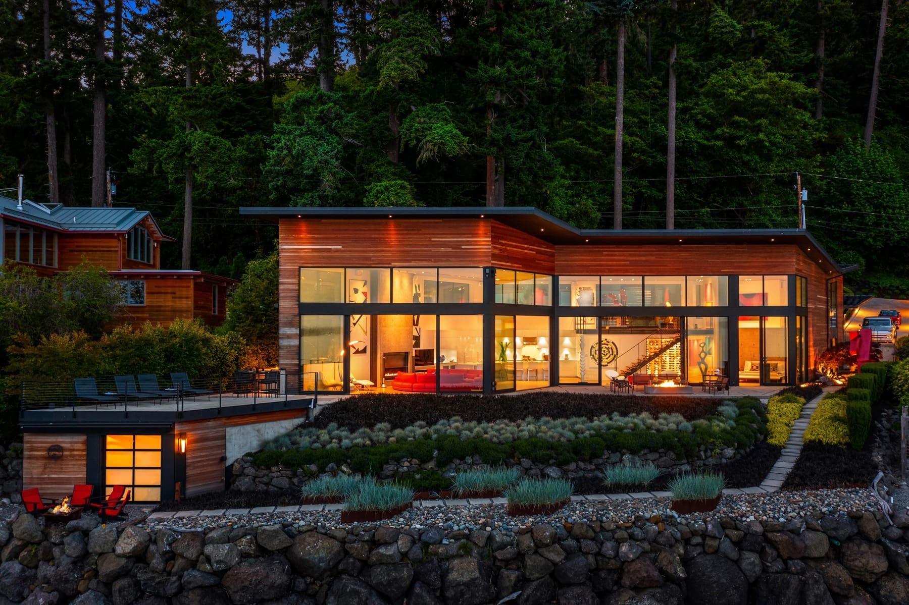 A modern, two-story house with large glass windows illuminated from the inside at dusk. The building features a sleek, angular design with wooden and metal elements. Surrounding the house are landscaped gardens, a patio with lounge chairs, and dense trees in the background.