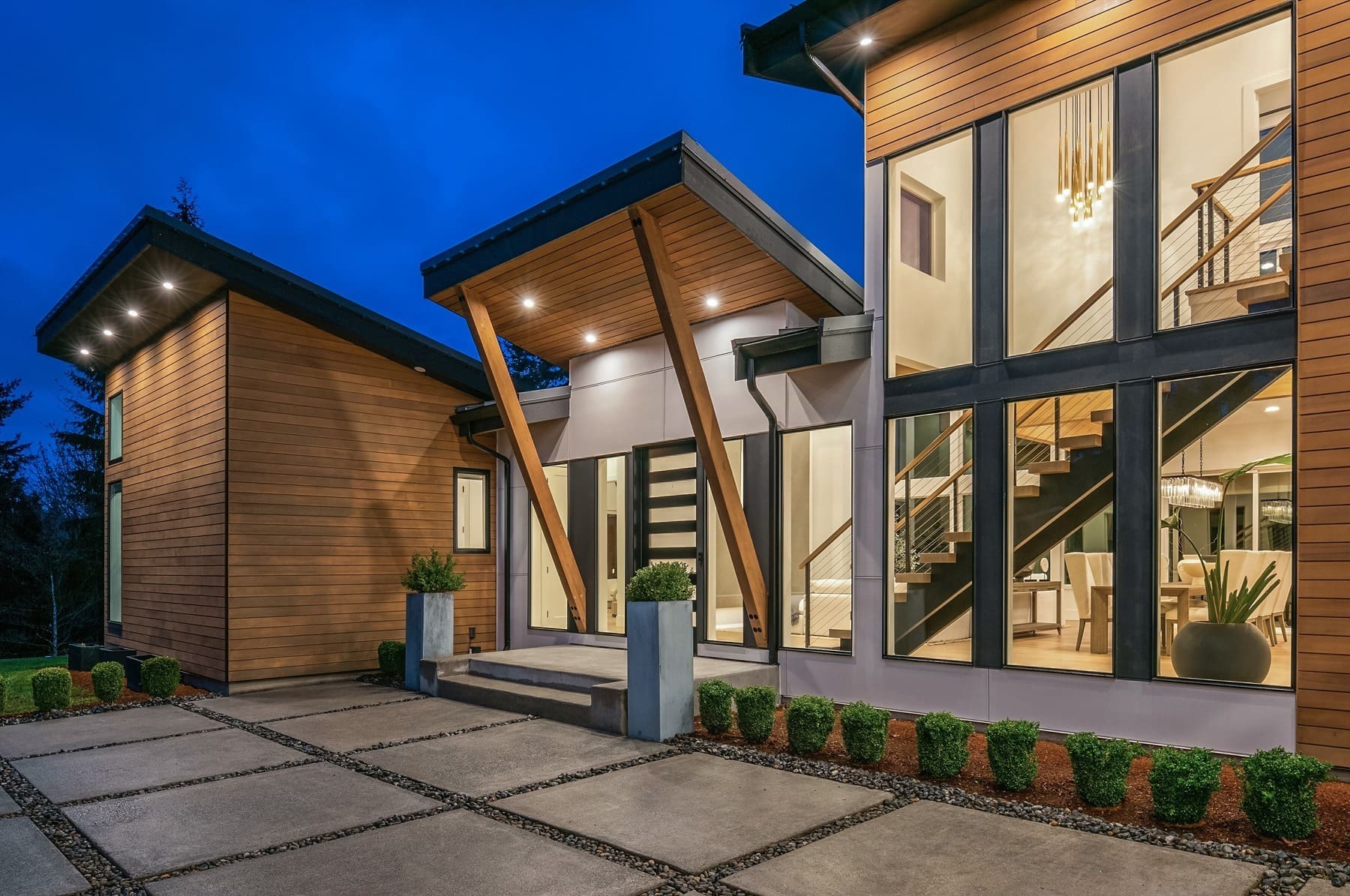 A modern house with prominent wooden paneling and large glass windows is illuminated at dusk. The front entrance features a sleek design with angular wooden beams and minimalist landscaping, including neatly trimmed shrubs and potted plants.