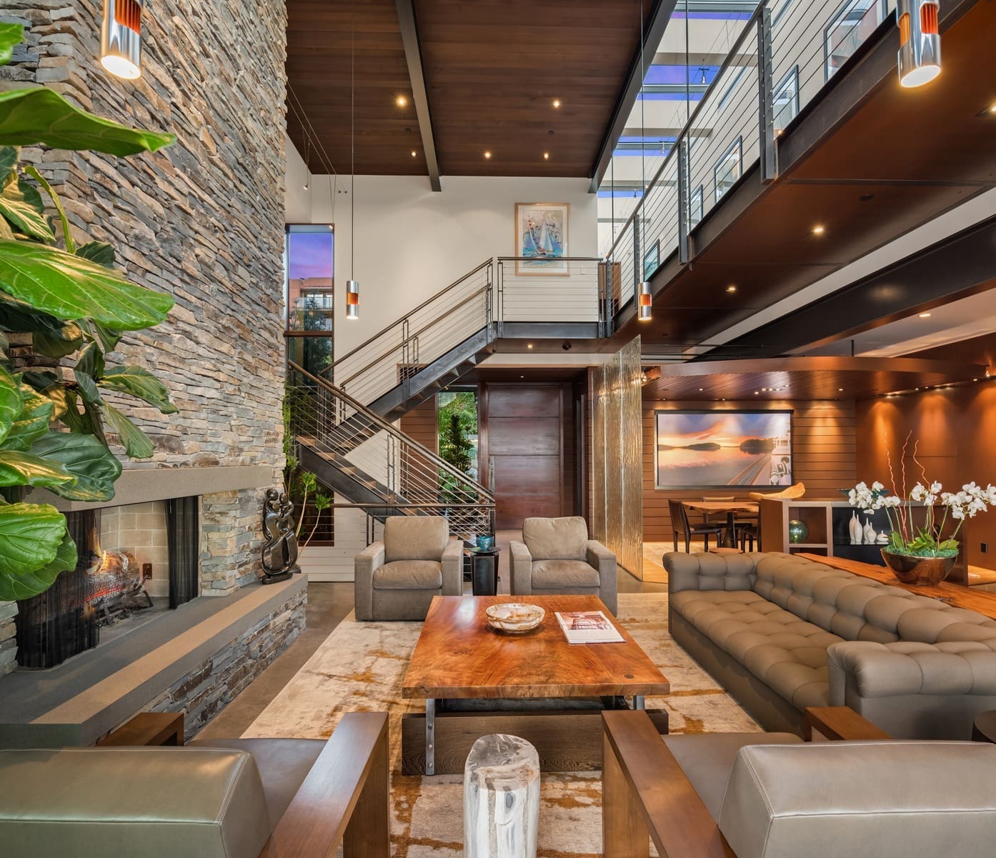 A modern living room with high ceilings and an open staircase. A stone fireplace is on the left, surrounded by cozy seating including a large sofa. The room is decorated with contemporary furniture, plants, and art, with large windows letting in ample natural light.