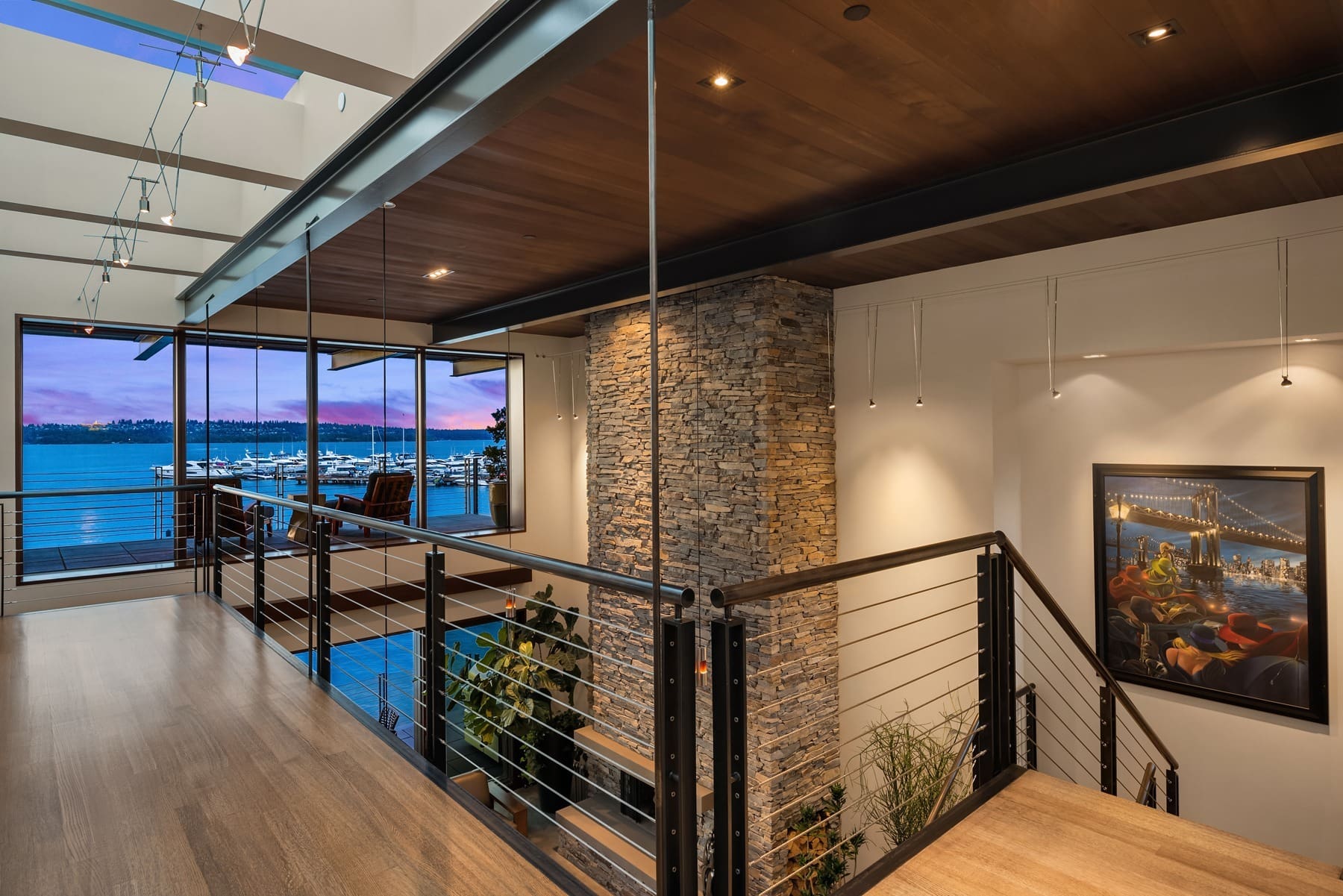 A modern interior with a wooden walkway, metal railings, and a stone accent wall. Large windows showcase a waterfront view with boats on water and a vibrant sunset. A painting featuring a cityscape hangs on the wall, and partially visible plants are seen below the walkway.