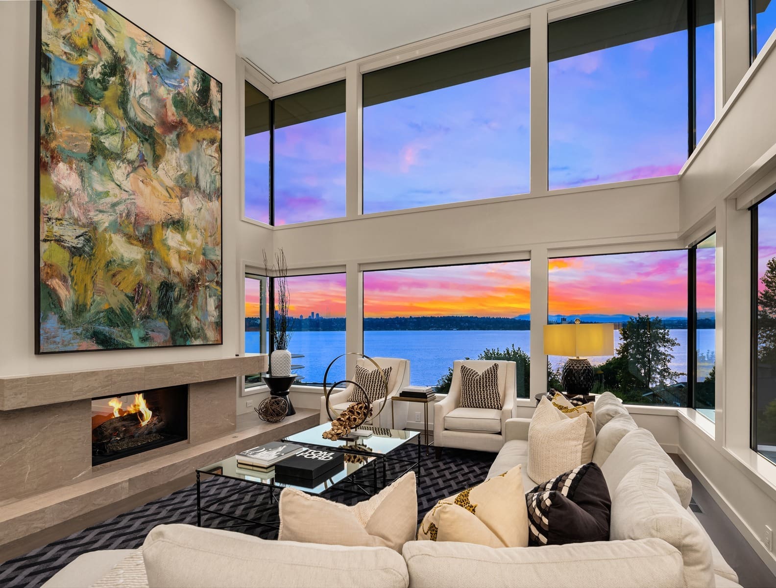 Twilight shot gallery1 A modern living room features floor-to-ceiling windows showcasing a stunning lake view and vibrant sunset. The room has a large sectional sofa with decorative pillows, a glass coffee table, a fireplace with abstract art above it, and stylish accents throughout. A charming, dark brown cottage with illuminated windows stands amidst a landscaped lawn at dusk. A small tree with a spotlight and an outdoor seating area enhance the cozy atmosphere. The clear sky overhead features a crescent moon, and tall trees frame the background. Twilight shot.