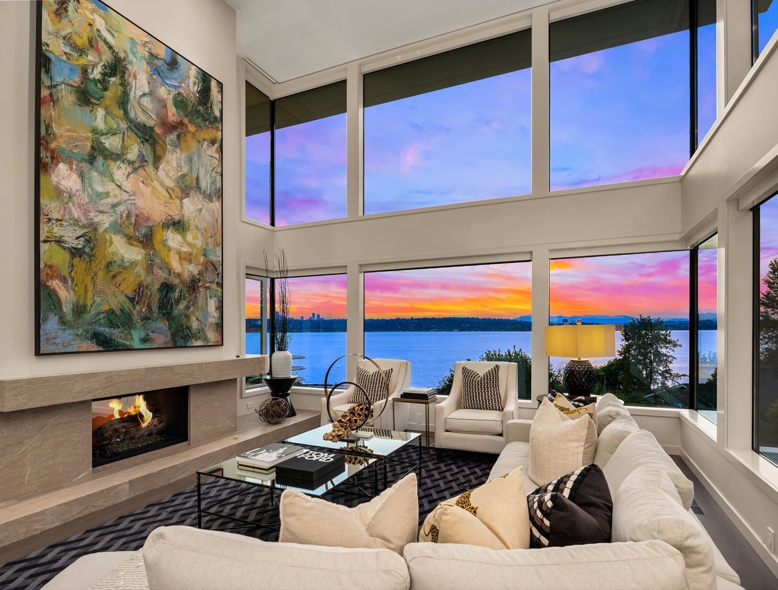 A modern living room features floor-to-ceiling windows showcasing a stunning lake view and vibrant sunset. The room has a large sectional sofa with decorative pillows, a glass coffee table, a fireplace with abstract art above it, and stylish accents throughout. A charming, dark brown cottage with illuminated windows stands amidst a landscaped lawn at dusk. A small tree with a spotlight and an outdoor seating area enhance the cozy atmosphere. The clear sky overhead features a crescent moon, and tall trees frame the background. Twilight shot.