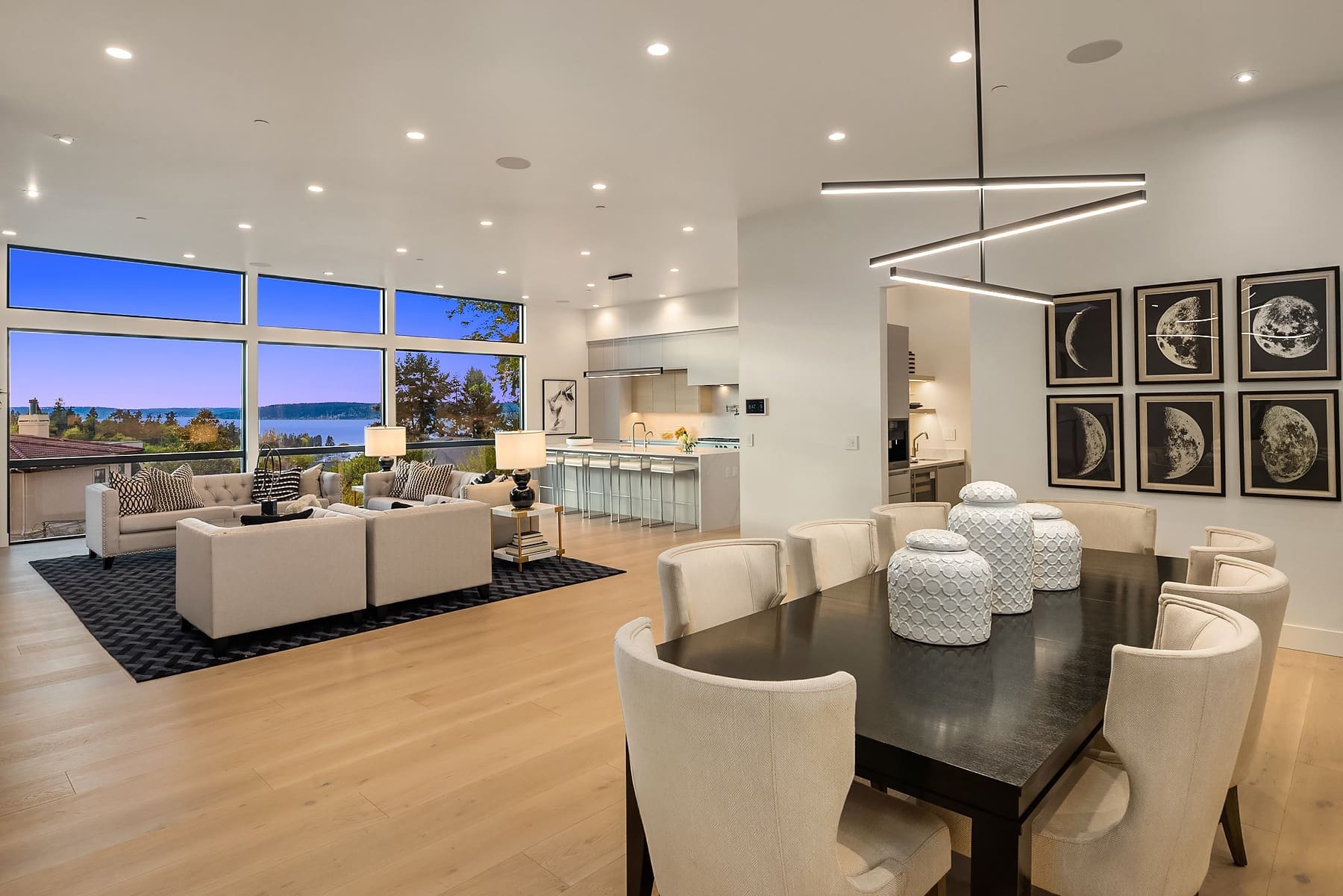 A modern, open-concept living space featuring a dining area with a dark table and white chairs, a spacious living area with light-colored sofas and a rug, and a sleek kitchen with bar stools at a counter. Large windows provide views of the outside scenery.