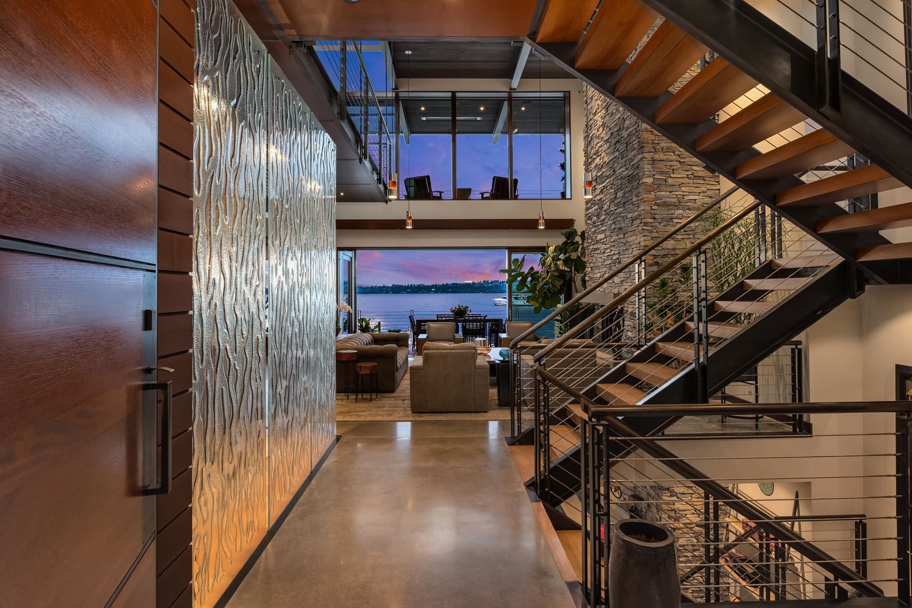 A contemporary home interior featuring an open-concept design with a view of a lake through large windows. The space includes a modern staircase, a stone wall, stylish furniture, and warm lighting. The polished concrete floor and wooden accents add to the aesthetic.