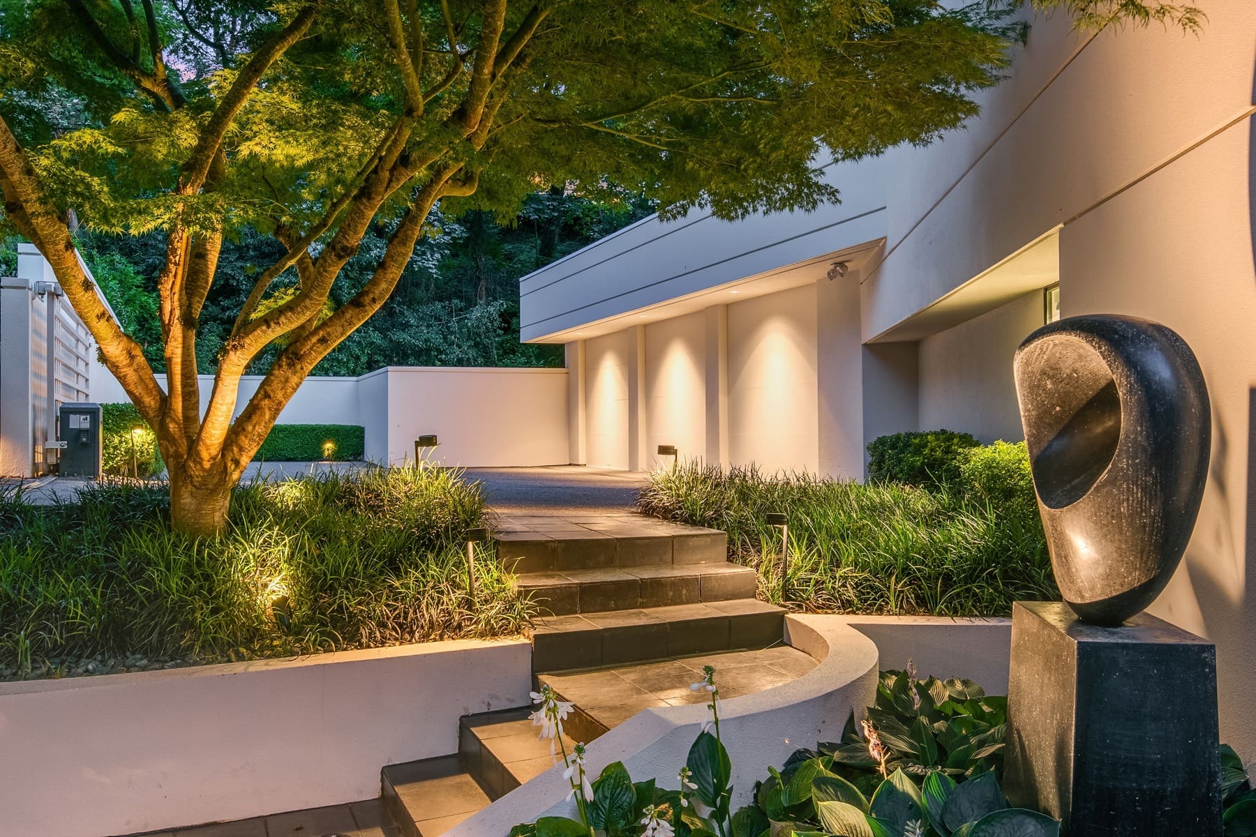 A modern house with a clean, minimalist exterior design. The entrance features a small flight of steps leading to a well-lit porch. A large leafy tree and sculptural artwork enhance the landscaped garden, adding to the serene atmosphere.