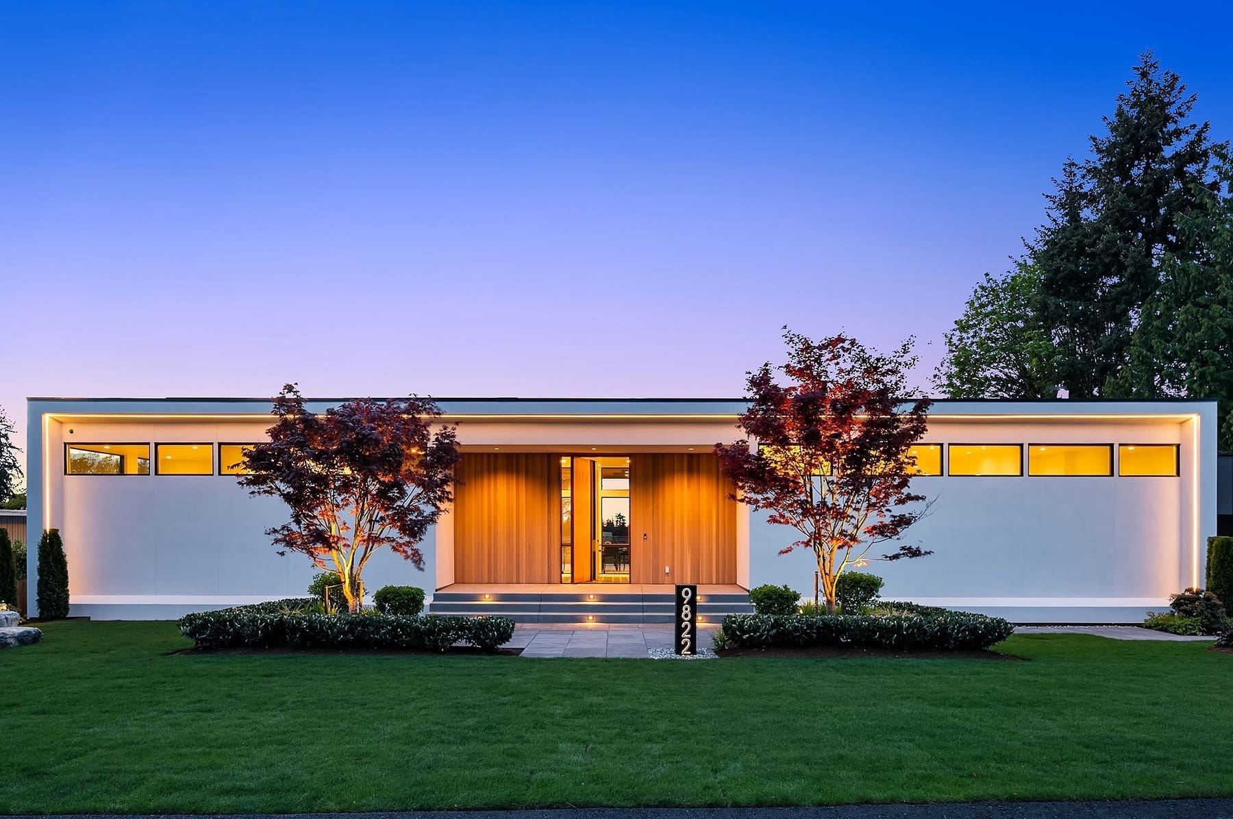 A modern, single-story house with large windows and a flat roof. The front yard features manicured grass, shrubs, and two trees. The house is illuminated from the inside, emitting a warm glow. A paved walkway leads to the front door, and the address is visible on a post.