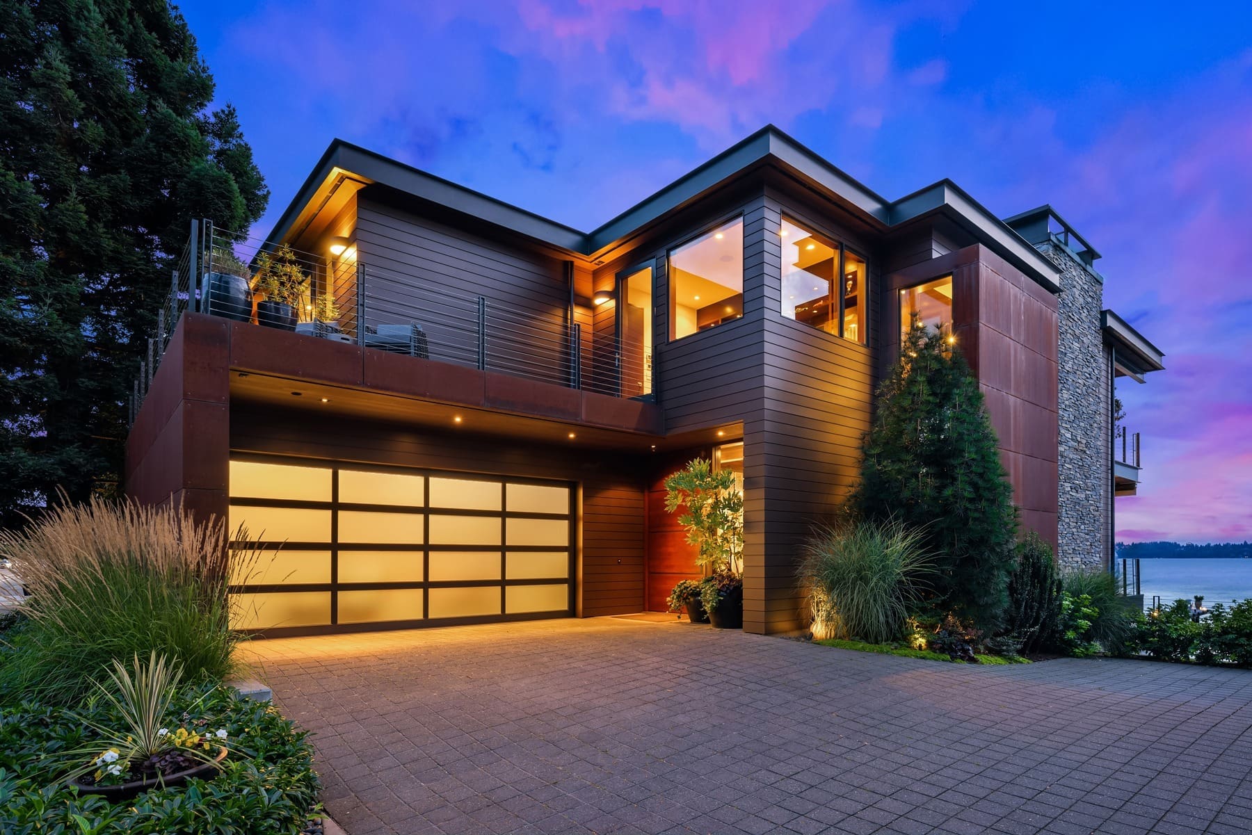 A modern two-story house with large windows, wooden paneling, and a glass-paneled garage door is illuminated by exterior lights during sunset. The well-manicured front yard features various plants and shrubs, and the lake is visible in the background. Twilight replacement.