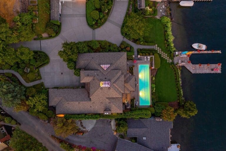 Aerial real estate photography view of a large house with a complex roof and well-maintained landscaping. The property features a swimming pool with a partially transparent cover, a driveway winding around the house, and a dock leading to a calm body of water with two boats moored.