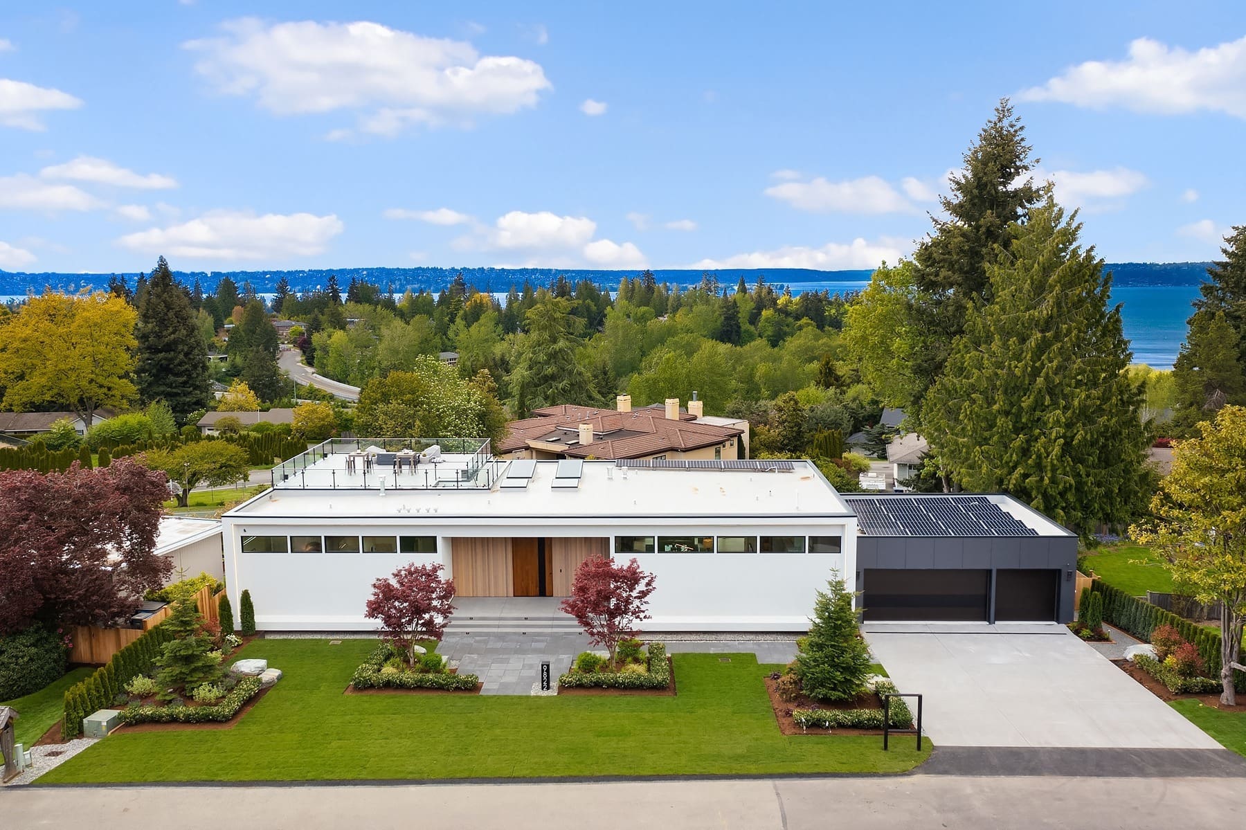 A modern, white rectangular house with a flat roof nestled in a suburban area. The property features a well-manicured lawn, two symmetrically planted trees, and a driveway leading to a black garage. The background reveals a lush landscape with tall trees and a distant lake.