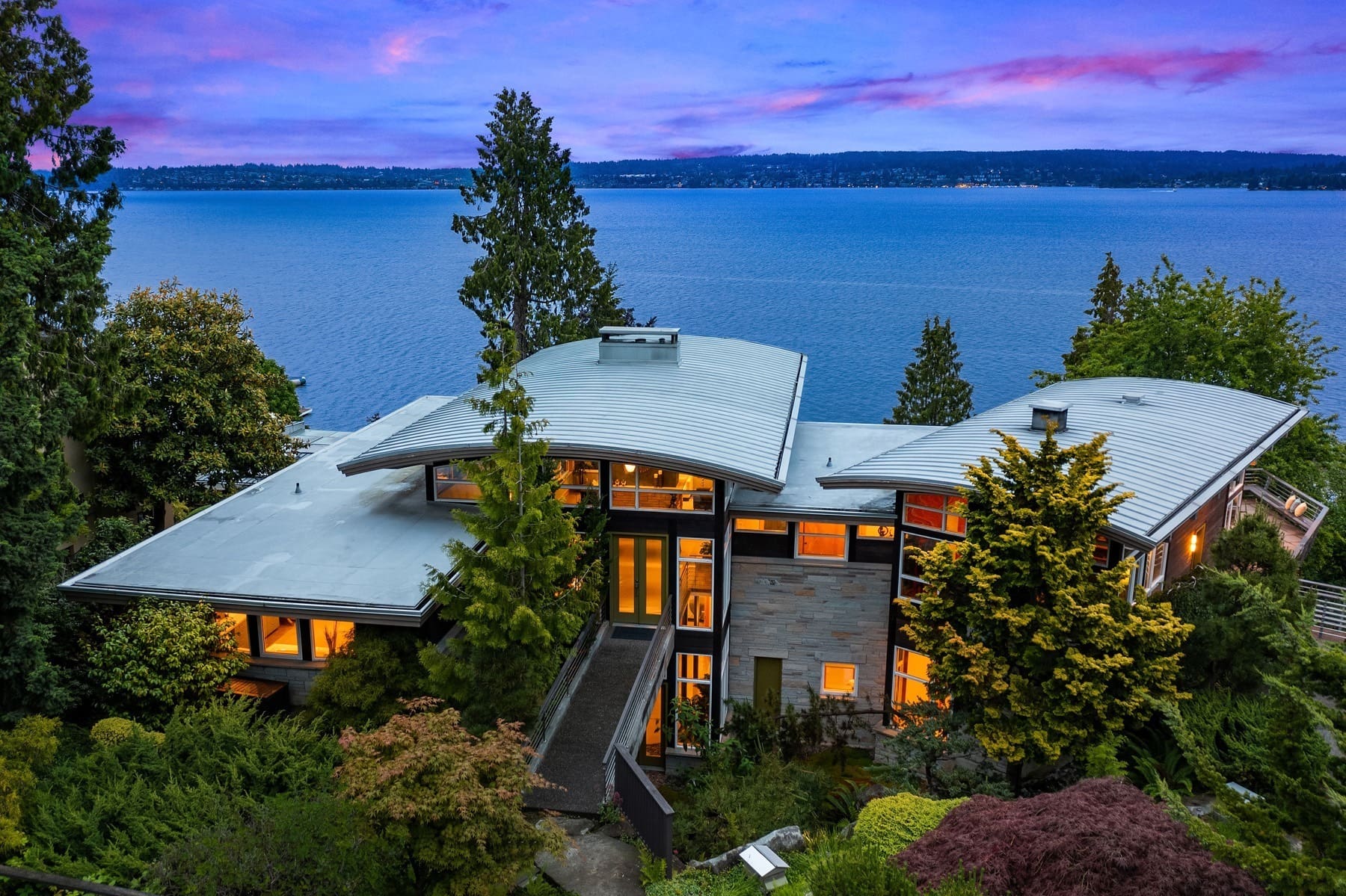 A modern, multi-story house with large windows sits surrounded by lush greenery. The home overlooks a large, tranquil body of water, with a vibrant, purple and pink sunset sky in the background, casting a serene glow on the scene.