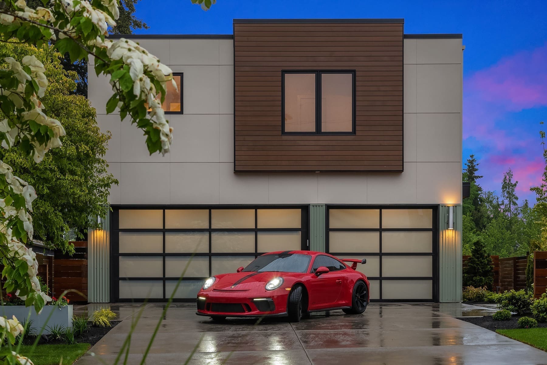 A modern two-story house with a sleek exterior design featuring large glass windows and wooden accents. Parked in front is a vibrant red sports car with black wheels, set against a backdrop of a vivid twilight sky. Lush greenery frames the driveway.