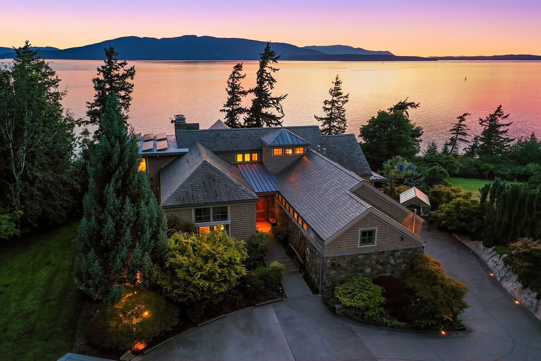 A luxurious house with a stone and wood exterior is nestled among trees on the edge of a body of water. The sun sets behind distant mountains, casting a colorful twilight sky. The property features a landscaped driveway and lush greenery.