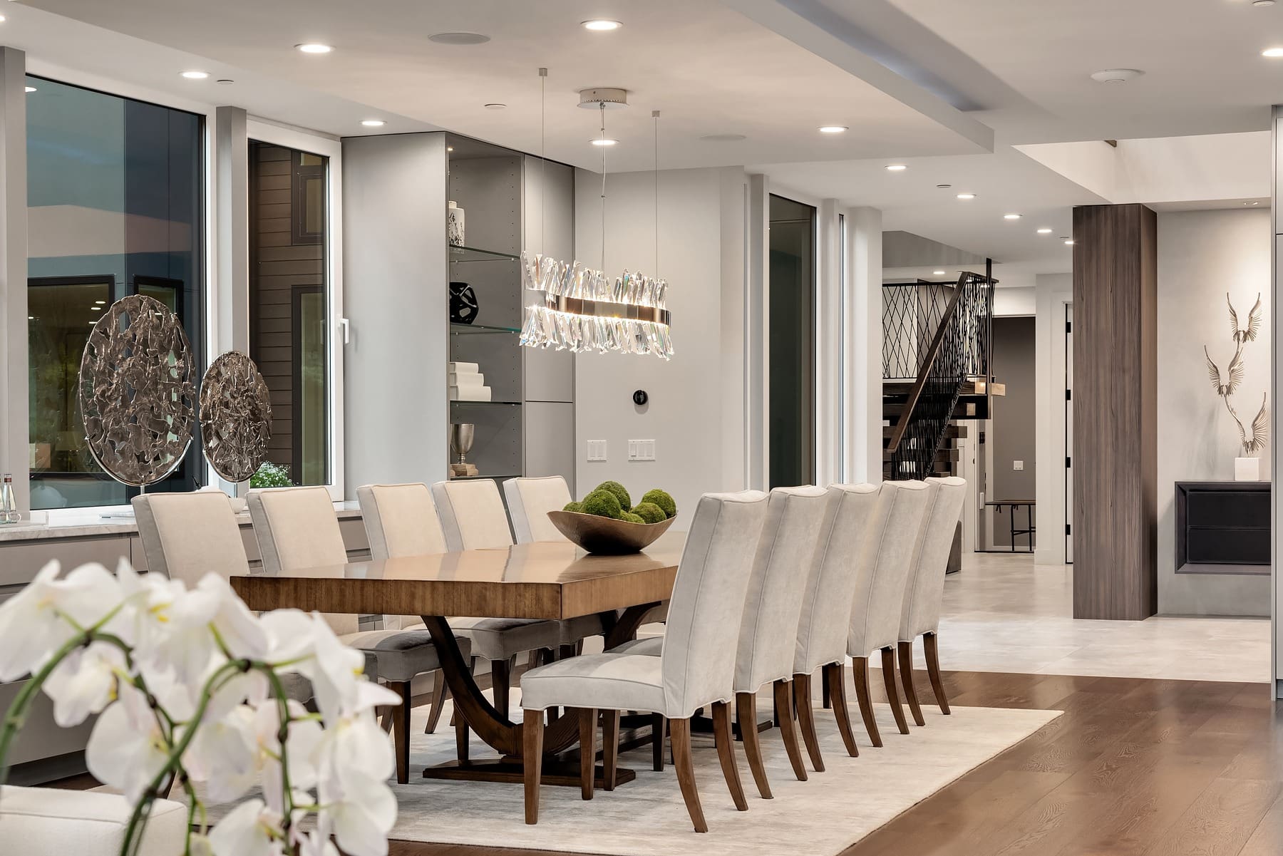 A modern dining room with a long wooden table surrounded by ten beige upholstered chairs. Contemporary light fixtures hang above the table. Large windows, decorative sculptures, and a staircase with a black railing are visible in the background.