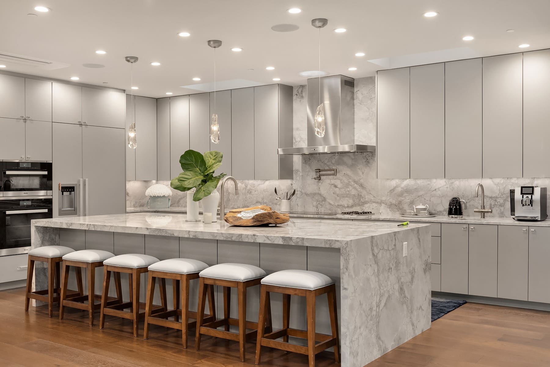 A modern kitchen features sleek gray cabinets, a marble island with four wooden barstools, and a stainless steel range hood. The island is decorated with a large plant and a loaf of bread. Pendant lights hang above, and under-cabinet lighting illuminates the workspace.