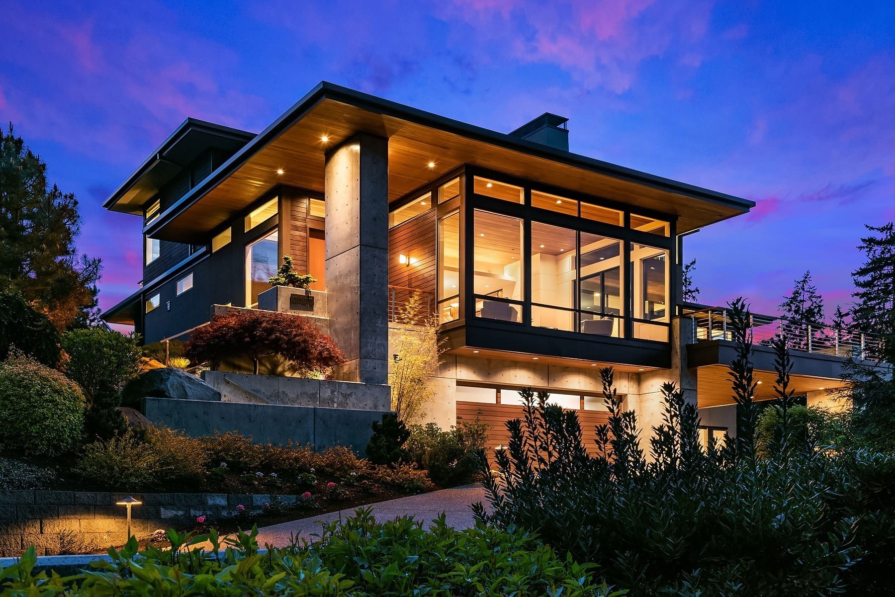 Modern two-story house illuminated at dusk with large glass windows and wooden accents. The home features contemporary architecture with clean lines and concrete elements. Surrounded by lush greenery and strategically placed outdoor lighting.