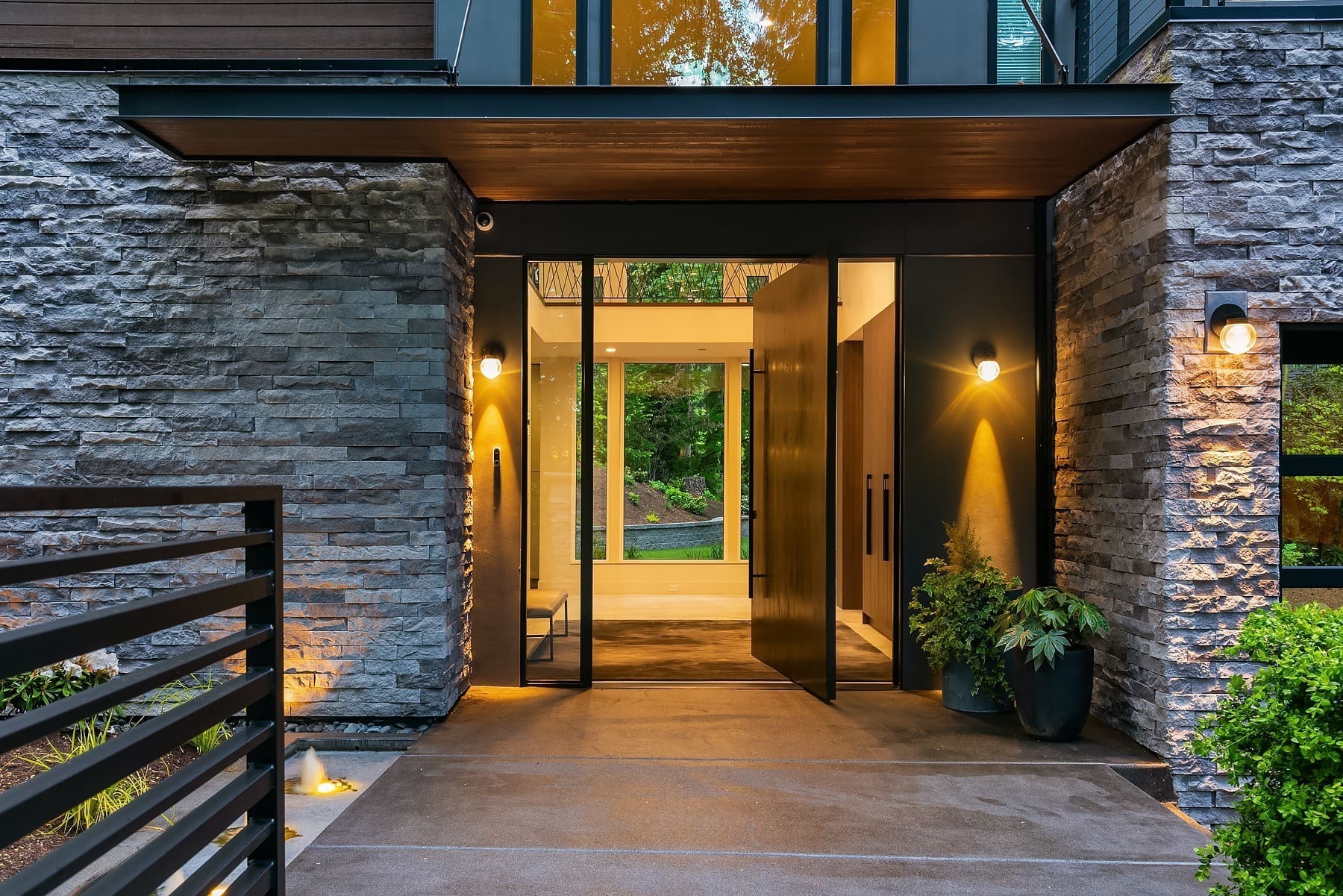 A modern house entrance with large double doors partially open, revealing a bright interior and garden view. The exterior walls are made of textured stone and smooth dark panels, with warm lighting and potted plants enhancing the inviting atmosphere.