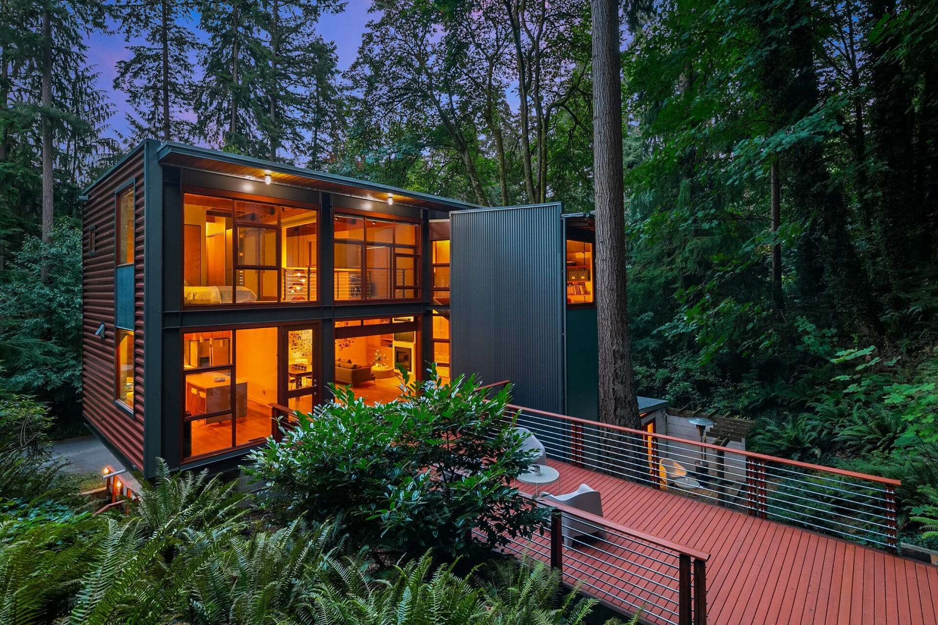 A modern two-story house with large glass windows is illuminated from within during twilight. Surrounded by dense forest, the home features a spacious deck with cable railing, integrating seamlessly into the lush greenery and towering trees.