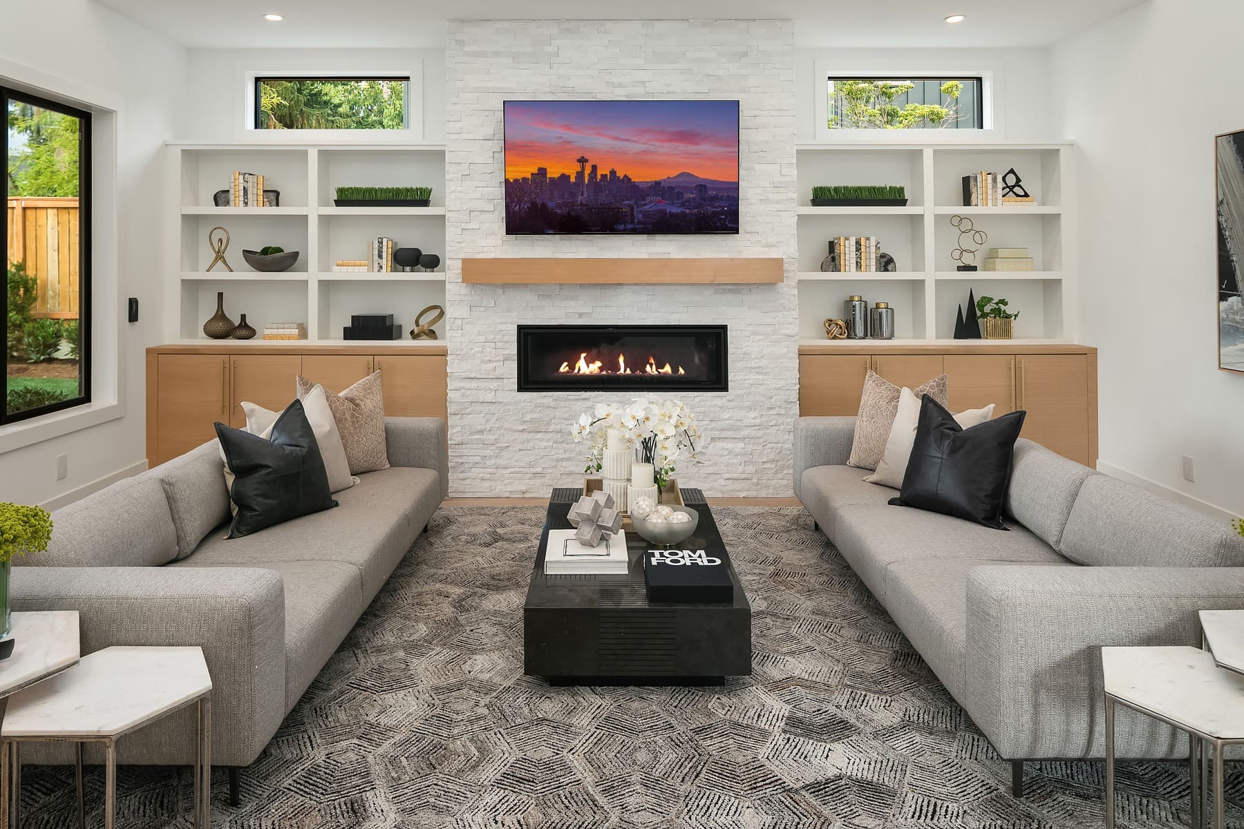 A modern living room with two light gray sofas facing each other, a black and white geometric rug, and a black coffee table with white orchid arrangement. The room features built-in shelves, a gas fireplace, and a wall-mounted television displaying a cityscape.