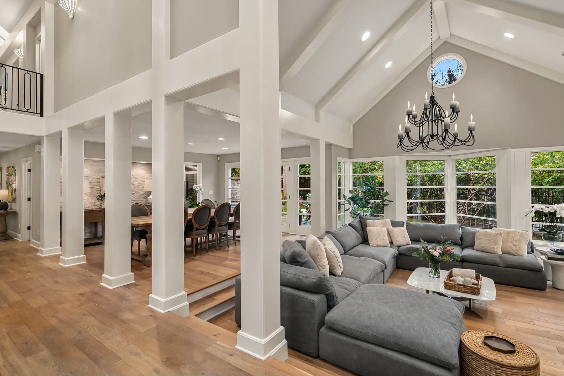 A spacious and modern living room with vaulted ceilings and large windows. The room features a grey sectional sofa, a chandelier, wood flooring, and plenty of natural light. Adjacent is a dining area with a table and chairs. Greenery is visible through the windows.