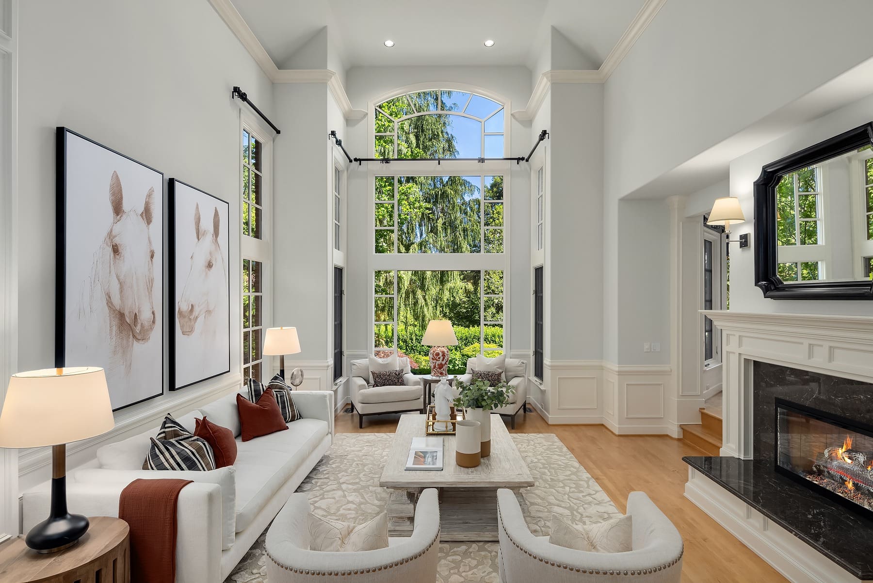 A spacious, bright living room featuring tall ceilings, large windows with a view of a lush garden, and abundant natural light. The room includes white couches, a modern coffee table, a cozy fireplace, and horse-themed artwork on the walls.