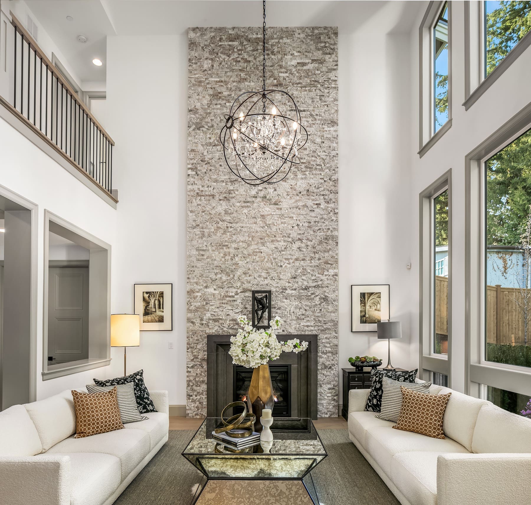 A modern living room with two white sofas facing each other and a glass coffee table in between. A tall stone fireplace extends to the ceiling, adorned with a chic chandelier. Large windows on both sides allow natural light to flood the space.