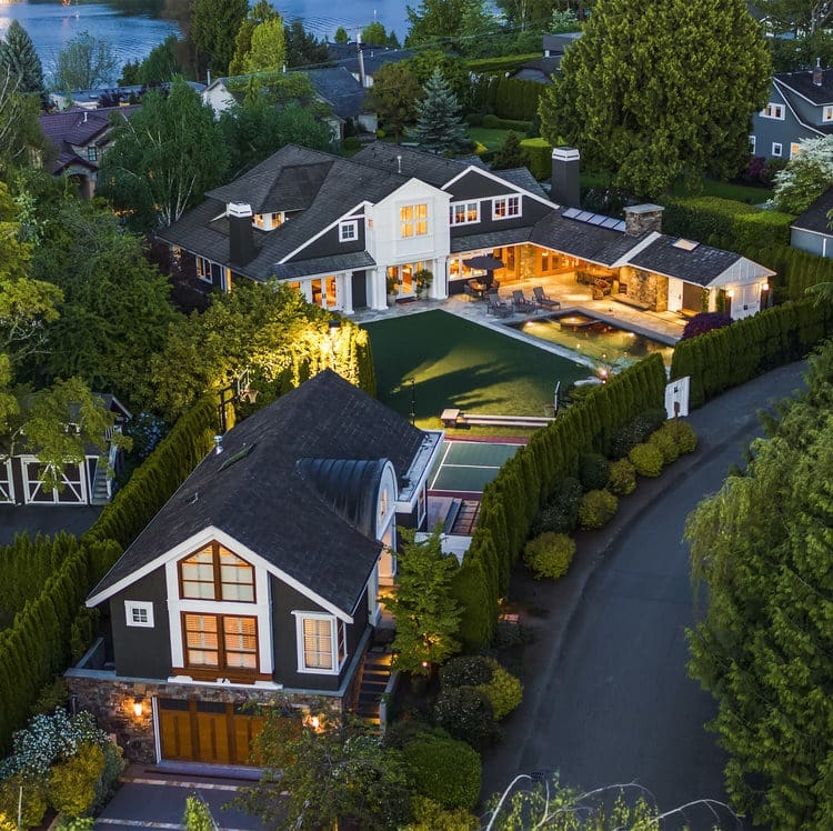 Aerial view of a luxurious property featuring two large houses with dark roofs and white trim, surrounded by dense greenery and landscaped gardens. The compound includes a pool area and overlooks a serene body of water in the background.