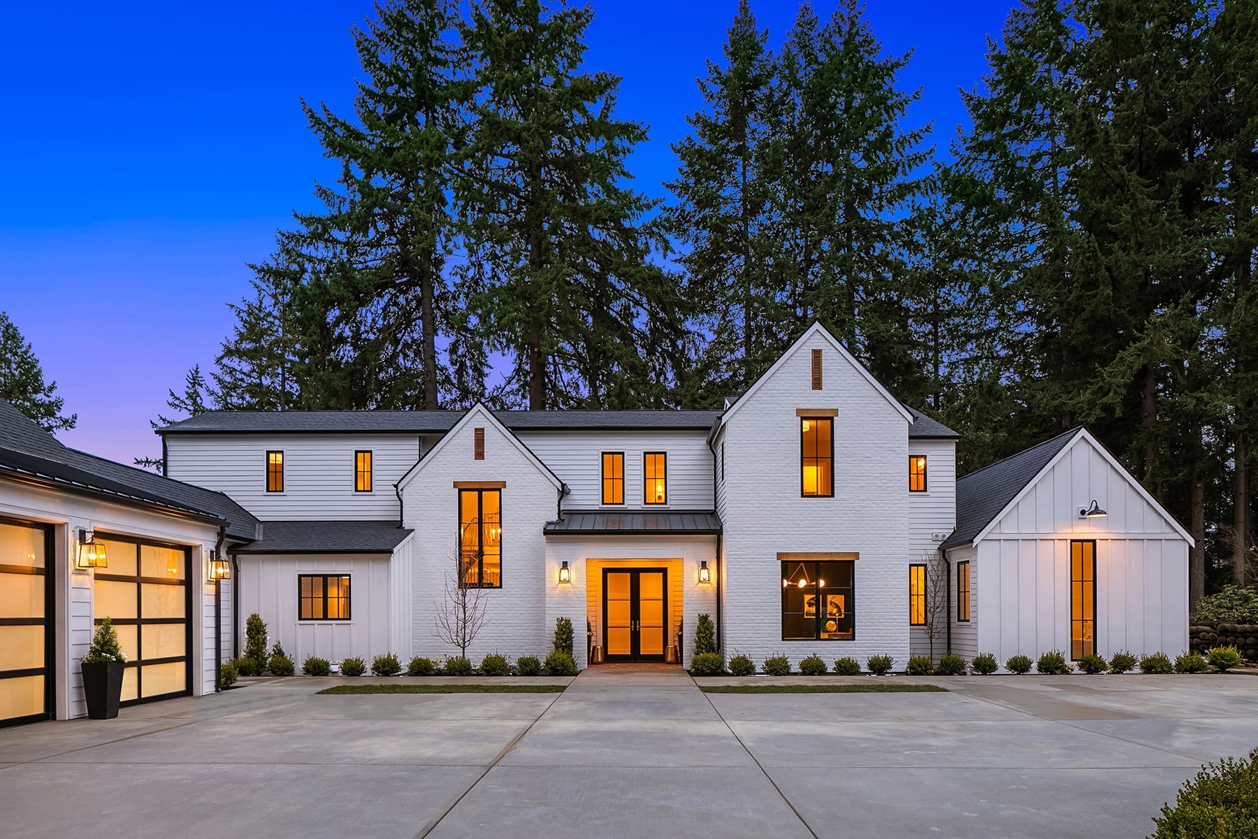 A large, modern two-story house with white brick exterior, large windows illuminated from inside, and a dark roof. The house is surrounded by tall trees and features a broad driveway with neatly trimmed shrubs lining the front. The sky is a clear twilight blue.