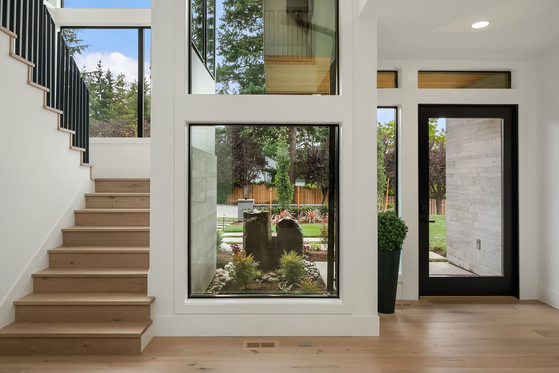 A modern home entryway featuring a wooden staircase on the left with black railings, a large window showcasing an outdoor garden in the center, and a black-framed glass door to the right. The interior is bright with natural light coming through the windows.