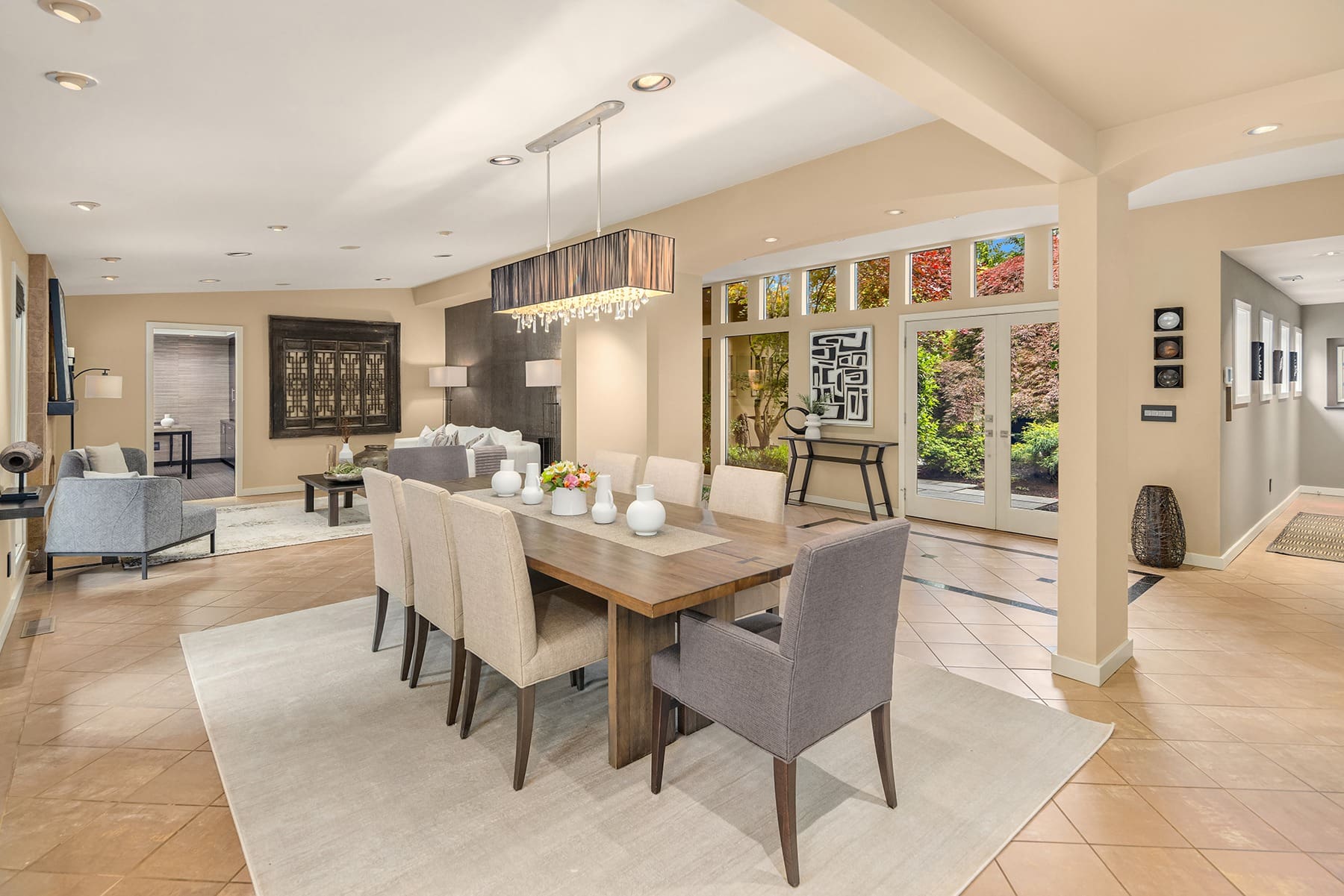 Spacious modern open-plan dining and living area featuring a wooden dining table with beige and gray chairs. A chandelier hangs above the table. The living area has a gray sofa, armchairs, and a coffee table. Large windows and glass doors offer views of a lush garden outside.