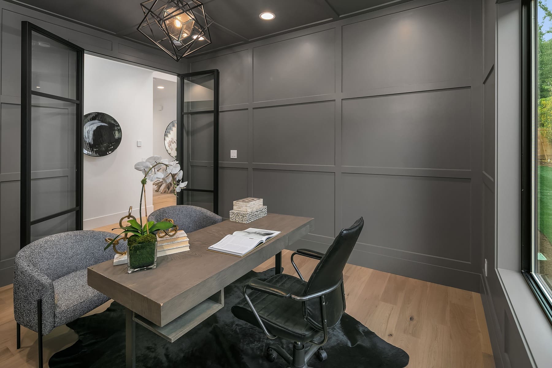 A modern home office with dark gray panel walls, a light wood floor, and a geometric light fixture. A wooden desk with a black office chair and gray upholstered guest chair sits on a black rug. The desk holds an open book, stacked papers, and a vase with an orchid.