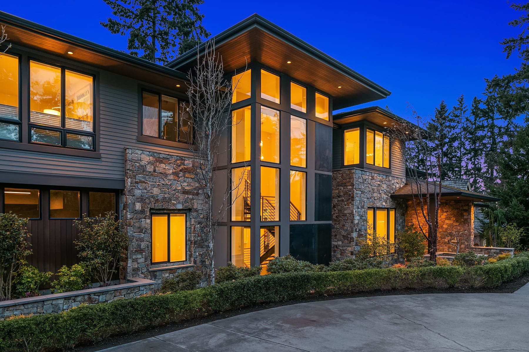 A modern two-story house, illuminated warmly from within, showcasing a glass-centered facade flanked by stone and wood paneling. The driveway curves gracefully in the foreground, framed by manicured shrubs. Tall trees surround the home at dusk.