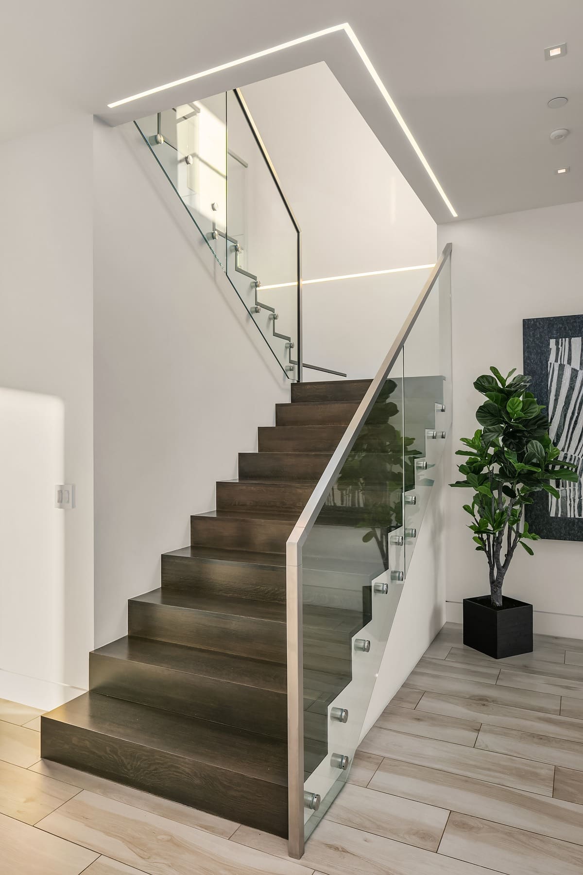 A modern interior staircase with dark wooden steps and glass railings, leading upward and turning to the right. The surroundings feature light-colored walls and a light wooden floor. A potted plant and a piece of abstract artwork decorate the space.