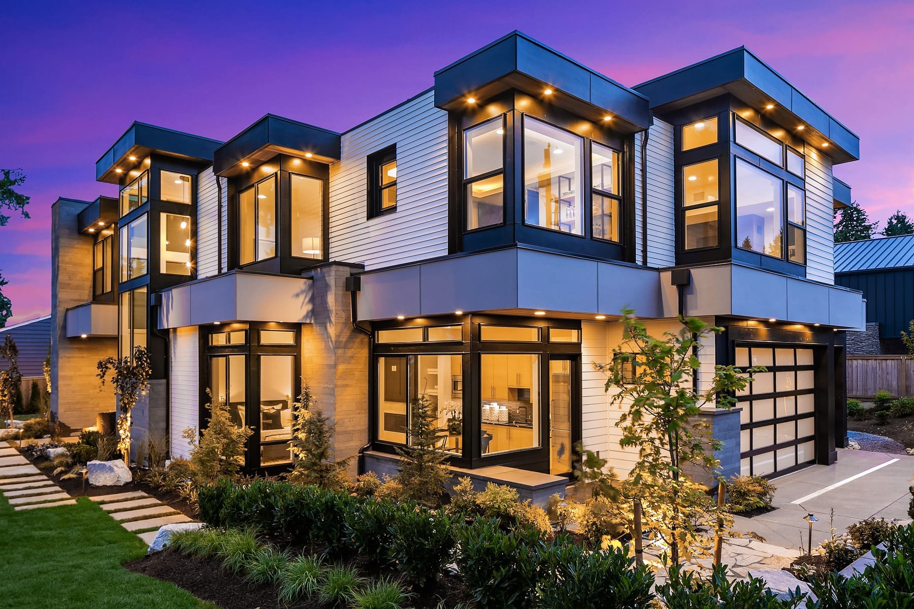 A modern, two-story house with large windows illuminated at dusk. The exterior features a mix of glass, metal, and wood accents. There is a surrounding garden with neatly trimmed shrubs and a pathway leading to the entrance. The sky is a deep twilight purple.