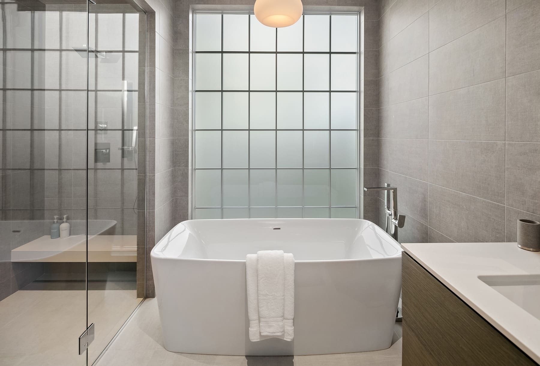 A modern bathroom with a white freestanding bathtub in the center. A towel hangs over the edge of the tub. There is a glass-walled shower on the left and a counter on the right. The walls are tiled, and a large frosted window allows natural light to fill the space.