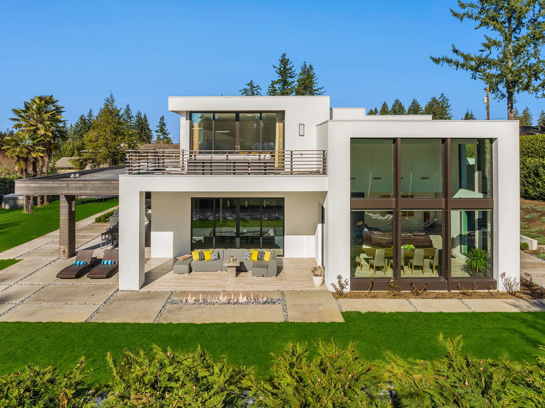 A modern, white two-story house with large glass windows and clean architectural lines. The spacious backyard includes a well-manicured lawn, outdoor seating area, and a pergola. The house is surrounded by trees and greenery.