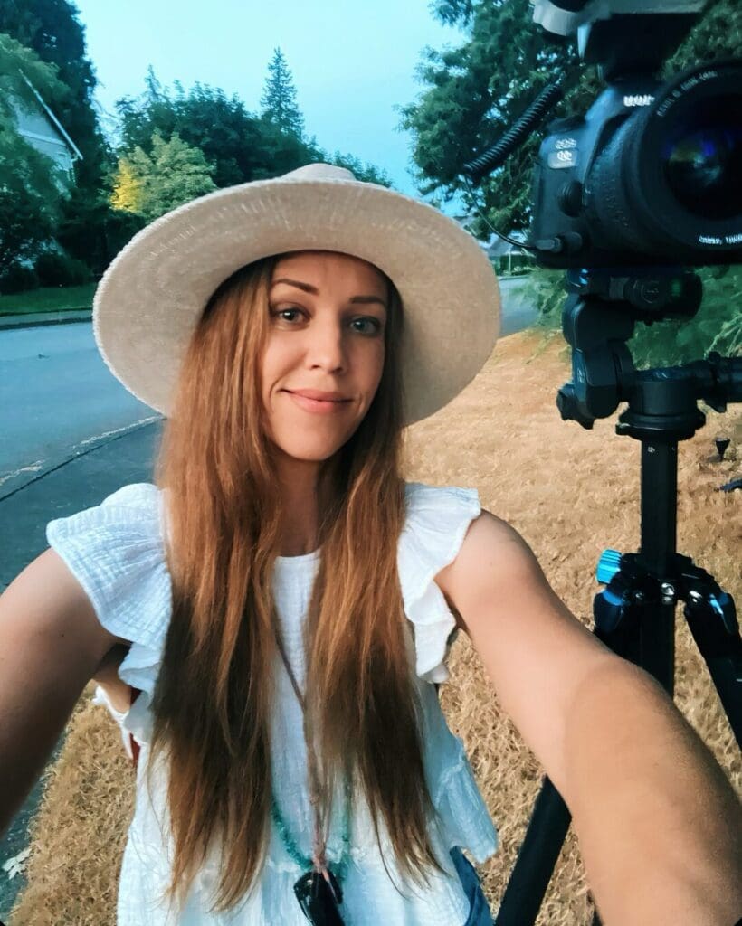 Self-portrait of a woman with long hair wearing a white ruffled top and a wide-brimmed hat, standing outdoors during twilight in a residential area, with a camera on a tripod nearby, showcasing the serene ambiance of the Pacific Northwest.