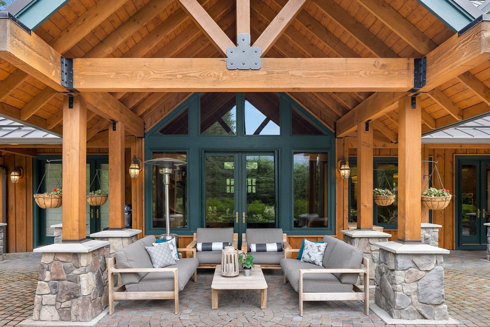 A covered outdoor seating area features wooden beams and stone pillars. Comfortable patio furniture, including two sofas and two chairs, encircles a wooden coffee table. Plants in hanging baskets and large windows are visible in the background.