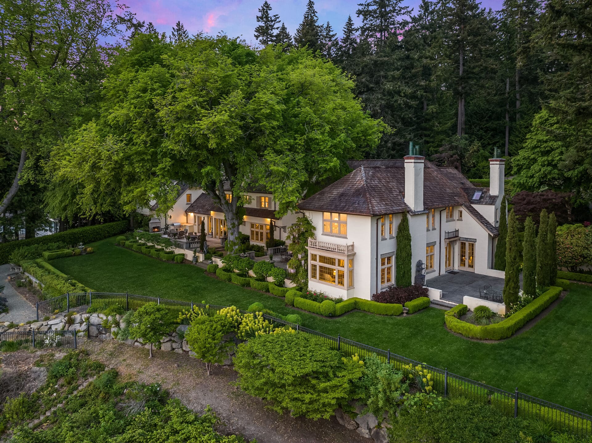 An aerial view of a large, elegant house with a spacious lawn and lush landscaping. The house has multiple chimneys, large windows, and is surrounded by dense trees and well-maintained hedges. A paved patio area with outdoor furniture is visible on the left side.