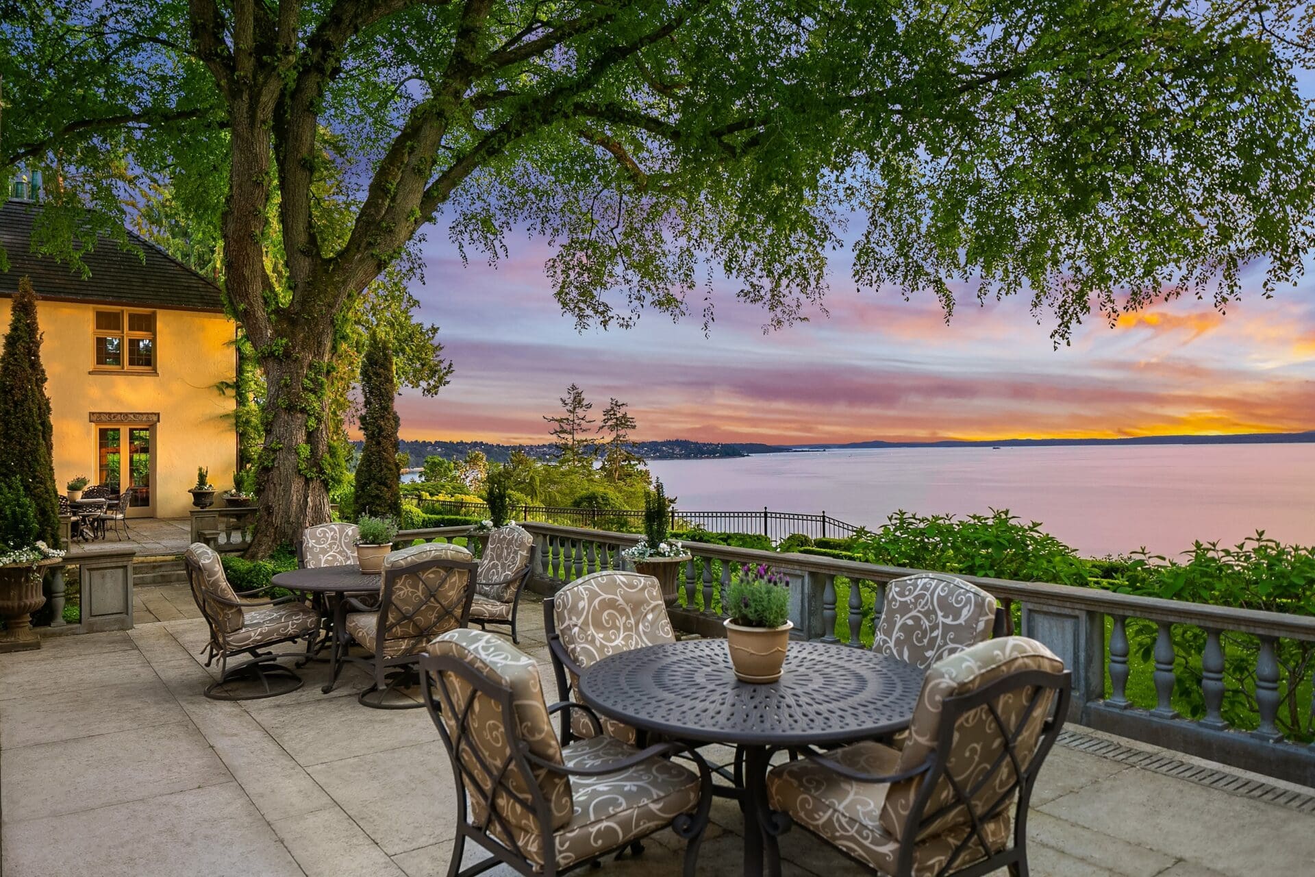 A scenic outdoor patio with elegant seating furniture under a large tree overlooks a serene body of water at sunset. The vibrant sky is a mix of orange, purple, and pink hues. A picturesque house with lit windows is on the left, bordered by lush greenery.