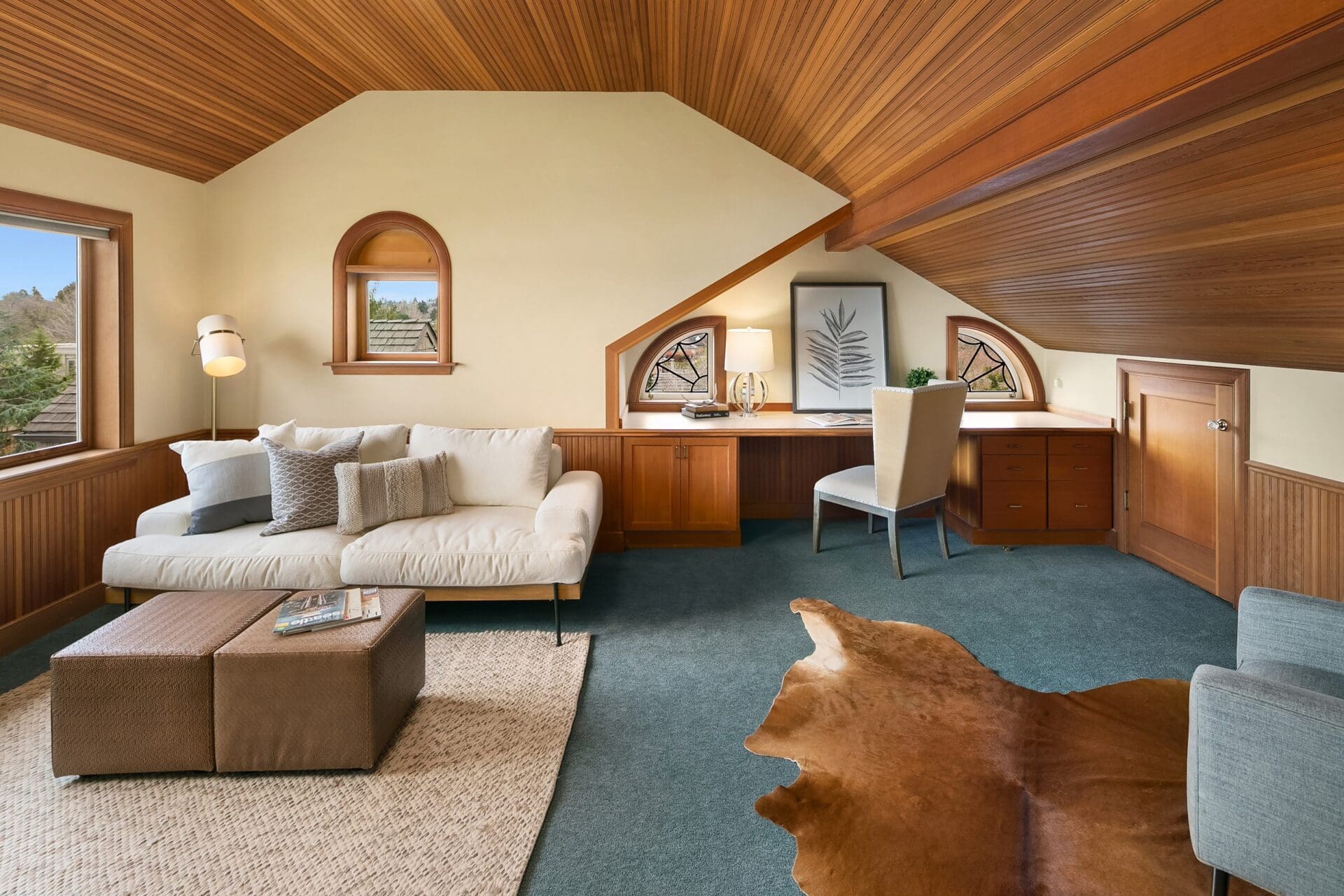 A cozy loft room with wooden ceiling and wall accents. It features a white sofa with beige and gray pillows, a brown ottoman, a woven rug, a desk with a chair, and windows providing natural light. There's a cowhide rug on the green carpet.