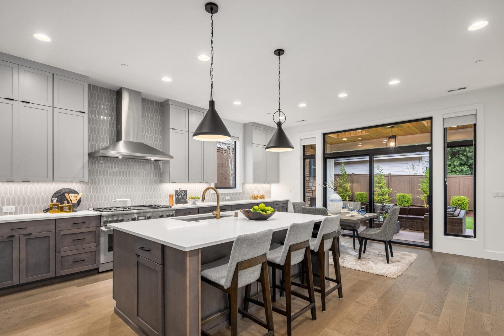 A modern kitchen featuring a large island with barstools and pendant lights above. It has stainless steel appliances, gray cabinetry, and a geometric tile backsplash. In the background, there's a dining area and sliding doors leading to a furnished patio outside.