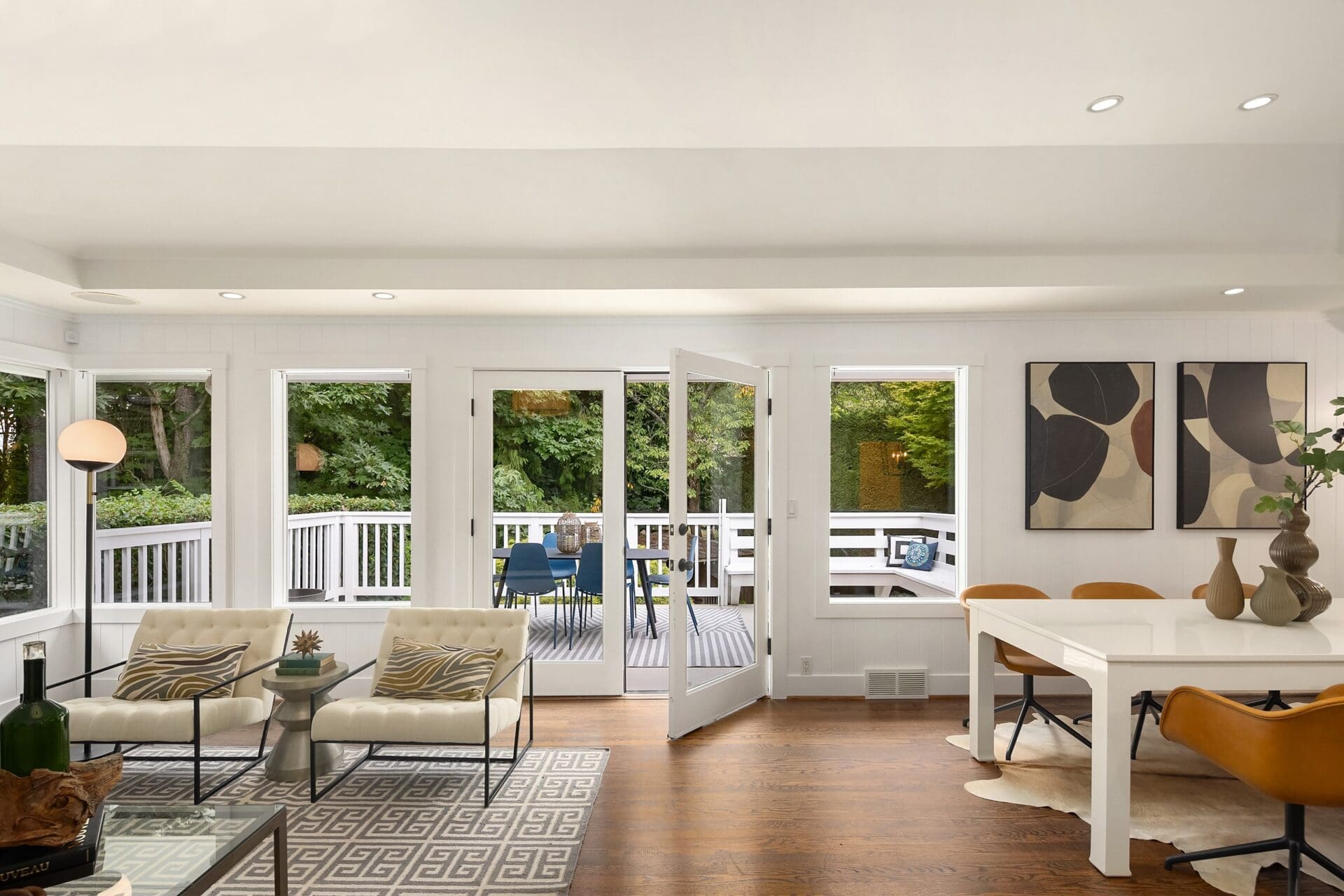 A modern living and dining area featuring a large glass door opening to a patio. The space has wooden floors, minimalist furniture, abstract art on the walls, and views of greenery outside. The decor includes neutral tones and contemporary design elements.