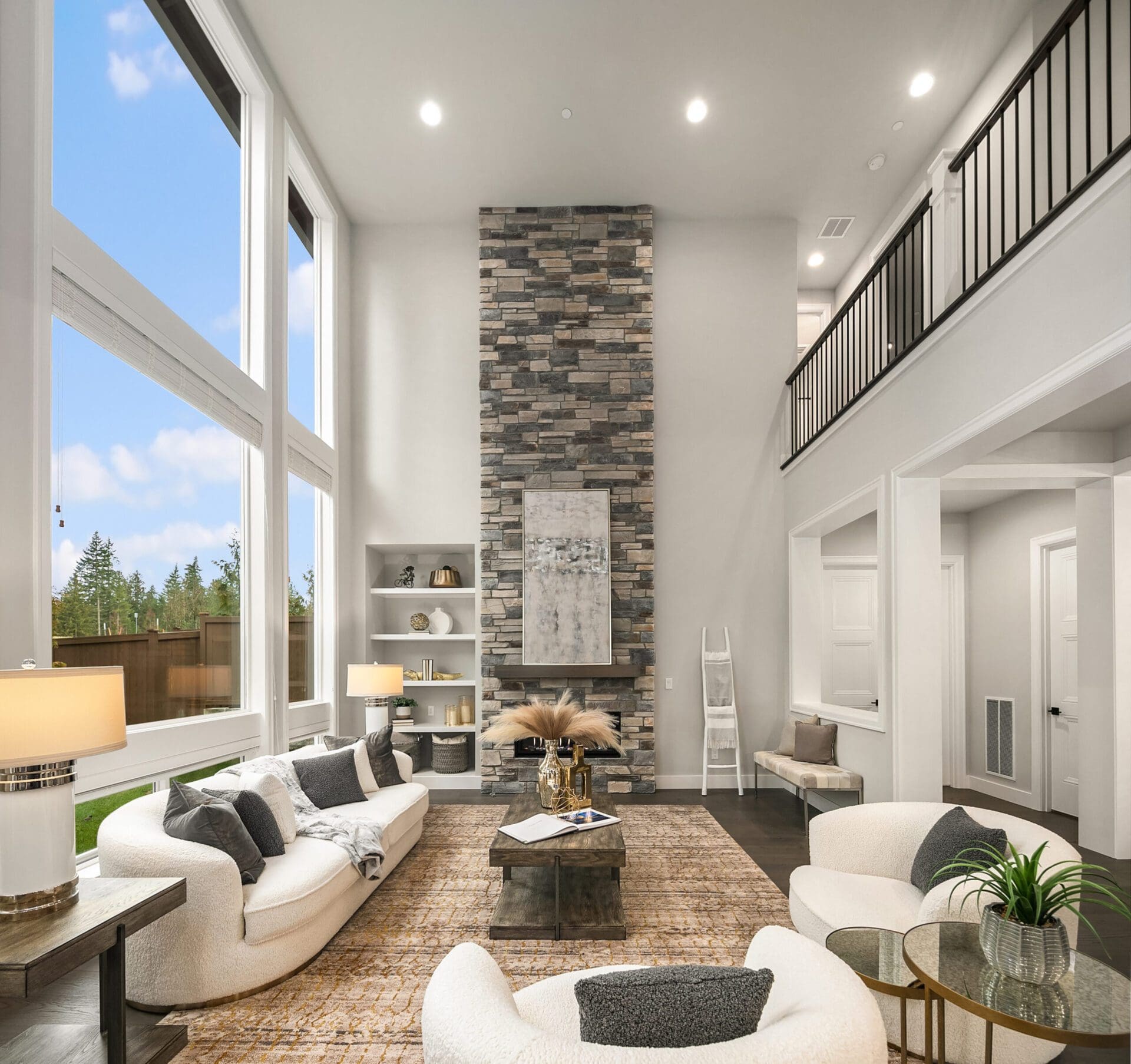 A spacious living room with high ceilings, large windows, and a floor-to-ceiling stone fireplace. The room features two white curved sofas, an armchair, a brown patterned rug, and a wooden coffee table with a decorative centerpiece. Shelves and decor pieces are along one wall.