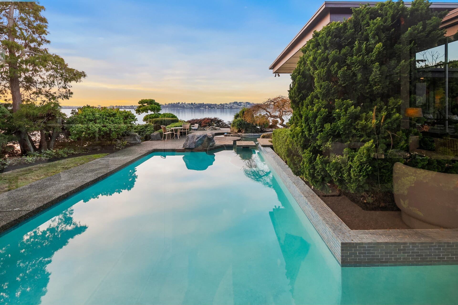 A serene backyard scene with a rectangular pool reflecting the clear sky and surrounding greenscapes. The pool is bordered by lush greenery and a modern house. Beyond the pool, distant trees and a body of water are visible, providing a tranquil atmosphere.